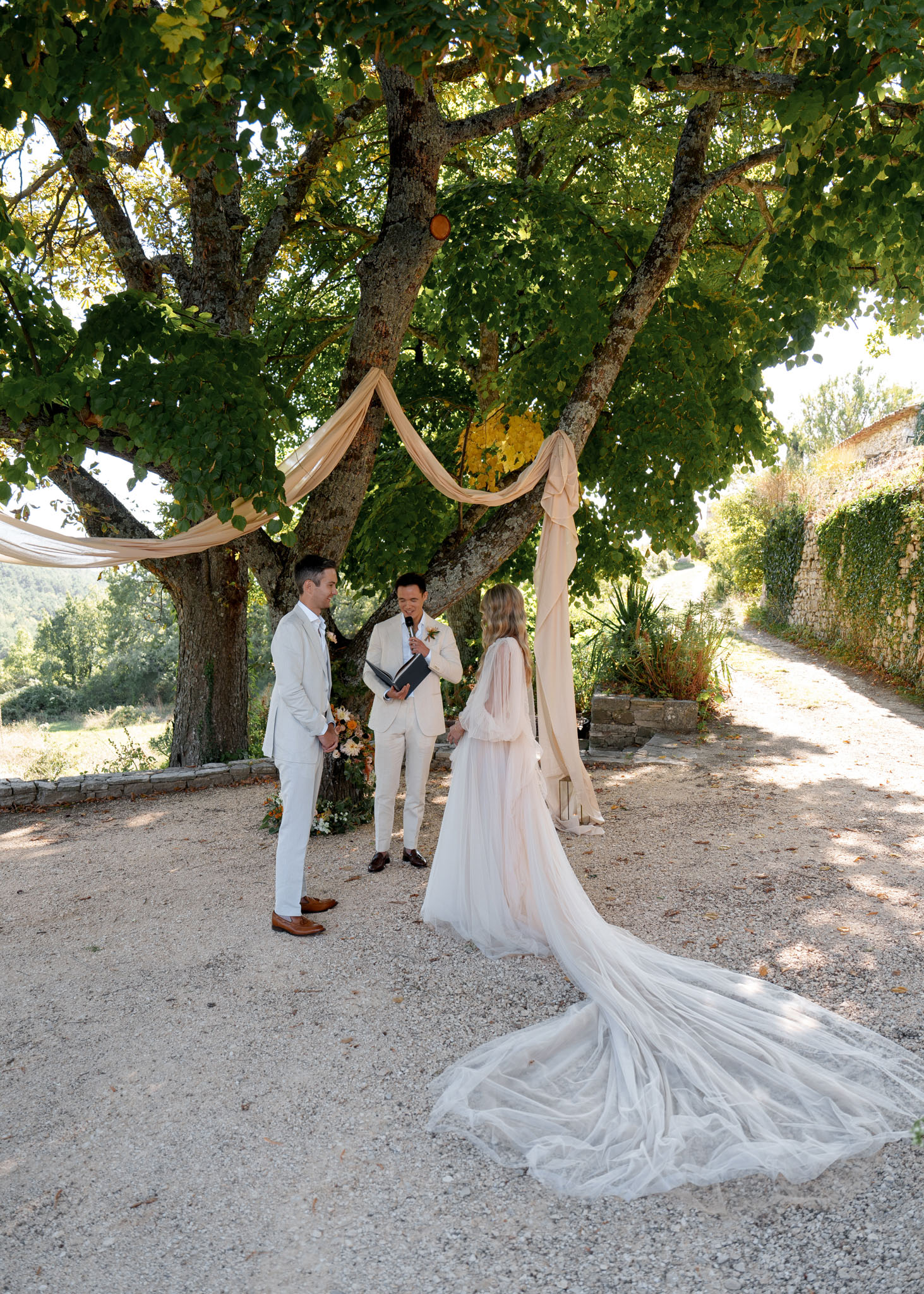 Outdoor ceremony under a tree with draped ivory fabric, bride in blush gown facing groom on gravel aisle
