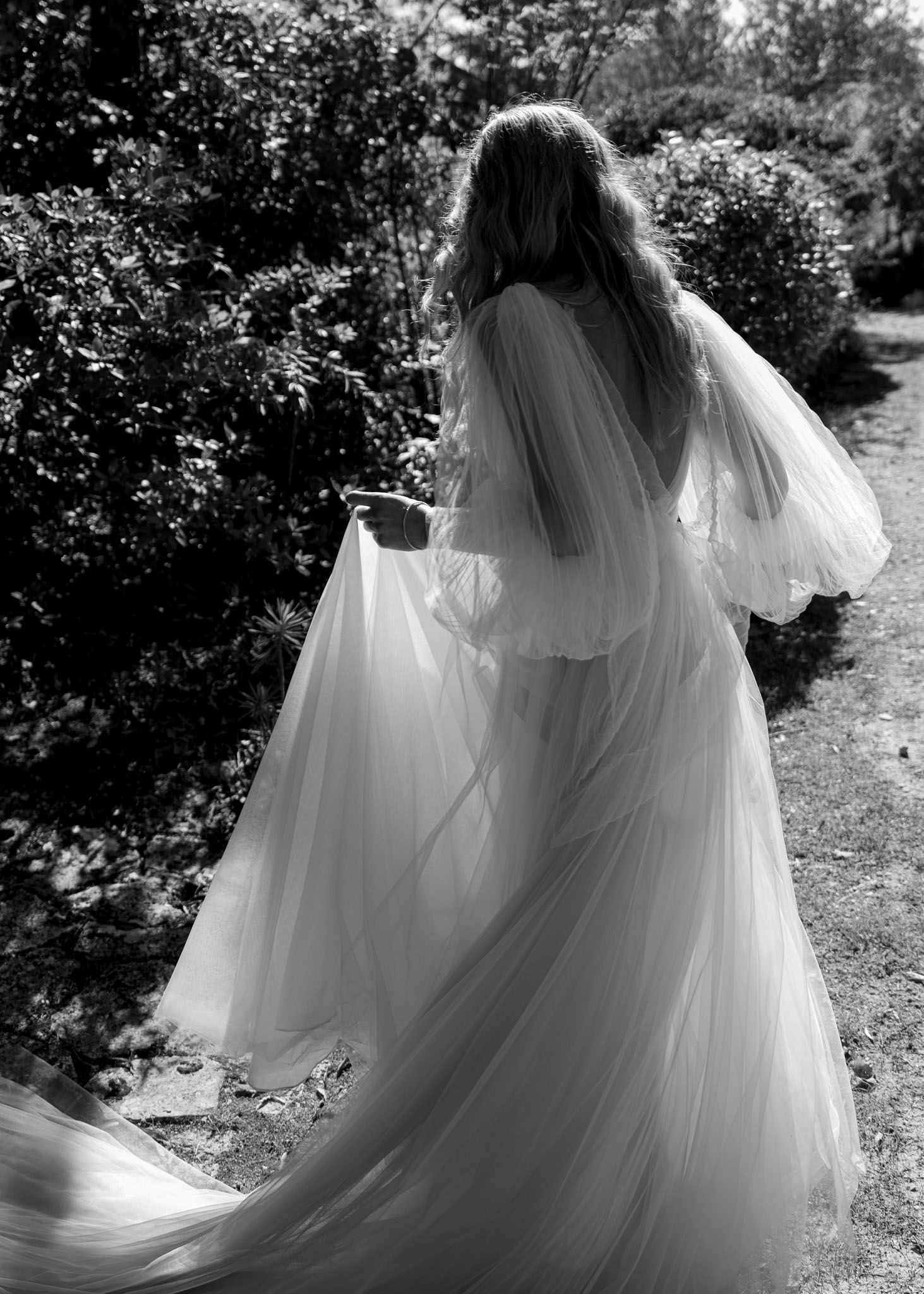 This black-and-white portrait shows a bride walking away from the camera along a garden path, her back to the lens. She wears a voluminous tulle ball gown with a low open back, dramatic puff sleeves with lace detailing, and a long cathedral-length train that fans across the ground. A veil layered over the dress adds further volume and movement. Her long wavy hair falls loosely down her back, and a bracelet is visible on her wrist. The high contrast between the bright white tulle and the deep dark tones of the surrounding garden foliage gives the image a strong graphic quality. The composition is a full-length rear portrait with the dress and train as the primary visual focus, suggesting a boho-romantic styling direction.