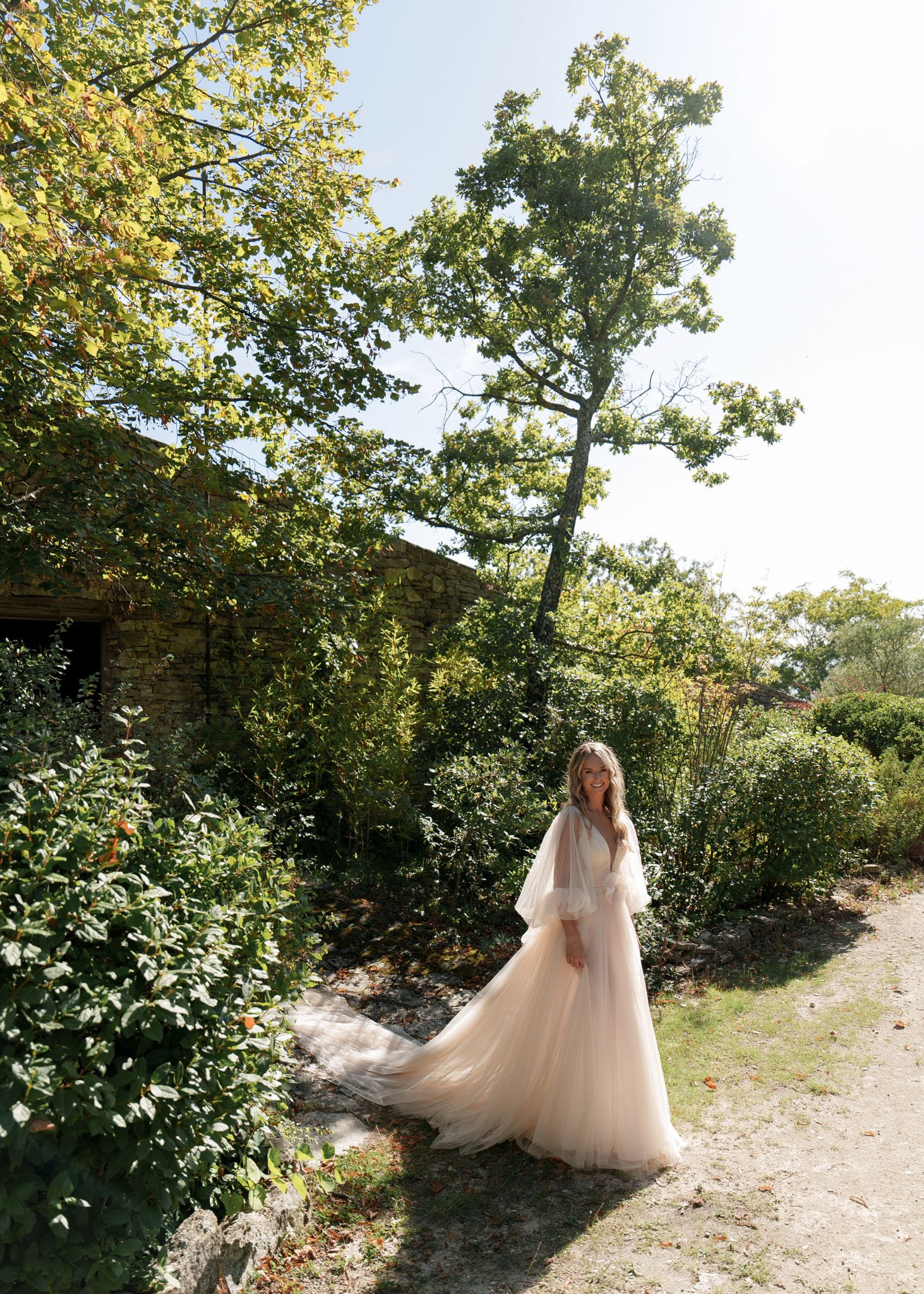 Bride standing on gravel path in blush tulle ball gown with flutter sleeves and long train beside stone building
