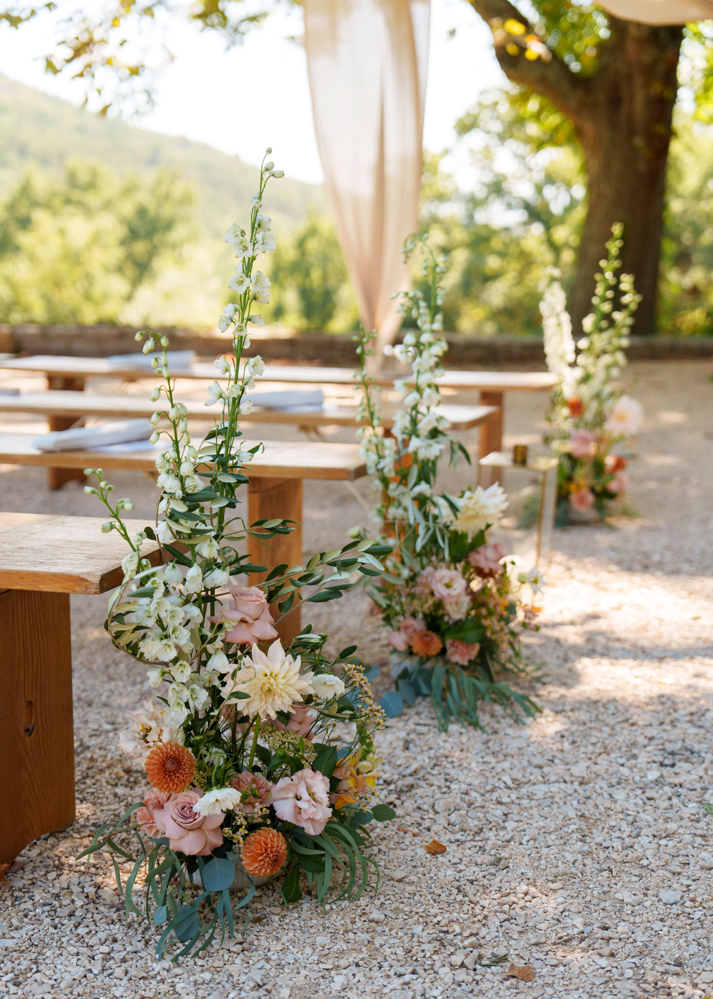 Detail shot of an outdoor ceremony setup on a gravel surface, showing aisle floral arrangements placed at the ends of wooden farm-style benches. The foreground arrangement features tall white delphiniums, cream dahlias, blush roses, orange and peach chrysanthemums or dahlias, pink lisianthus, and eucalyptus foliage. A second matching arrangement is visible in the mid-ground along the aisle, with a third partially visible further back. Ceremony programs or cushions are laid on the wooden benches. A draped cream fabric structure, likely an arbor or canopy, is partially visible in the upper background. The floral palette combines white, cream, blush, dusty rose, and burnt orange tones with green foliage, consistent with a garden-inspired or romantic rustic styling theme.