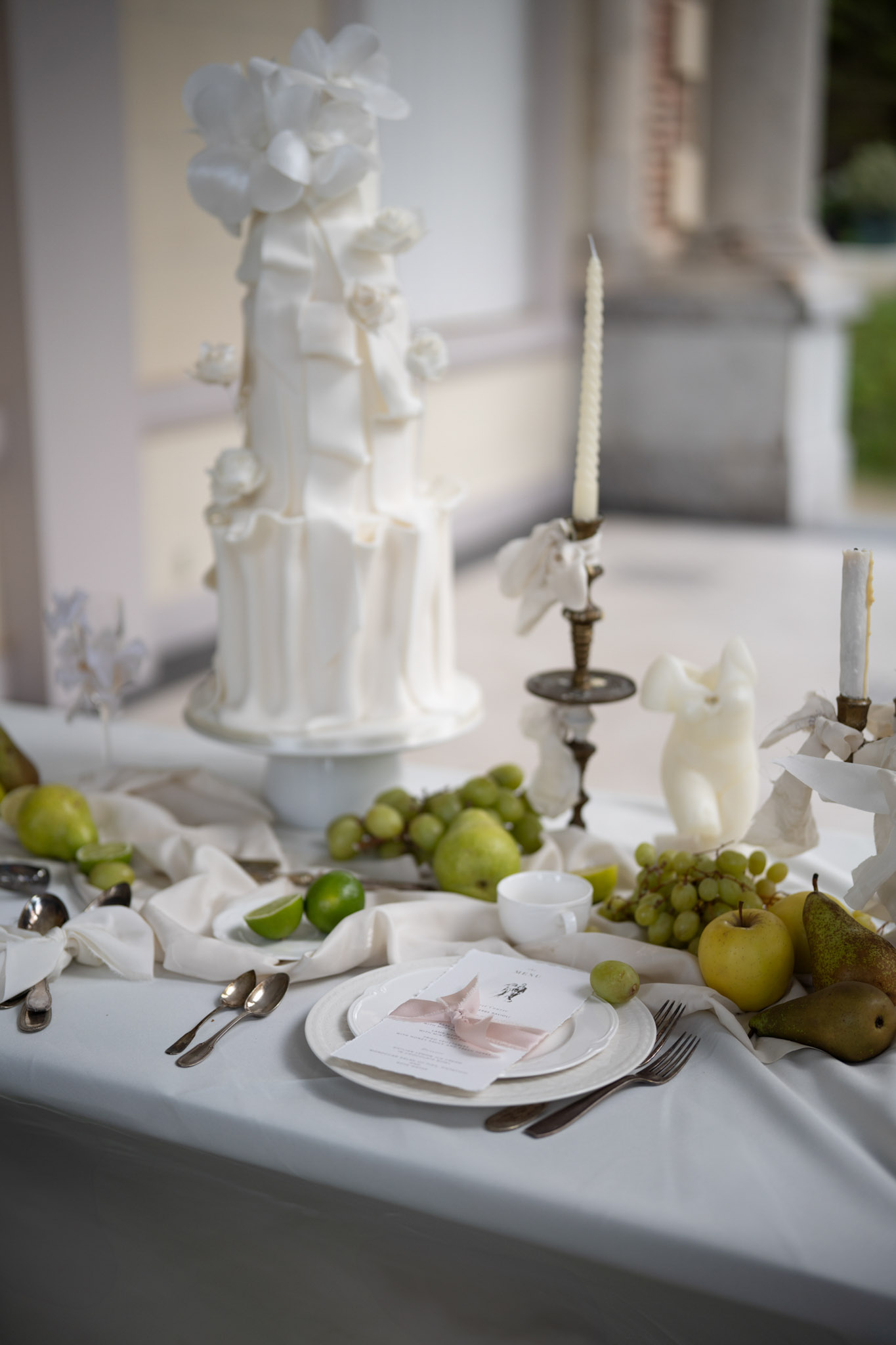 Three-tiered white wedding cake with sugar orchids beside tablescape with green fruit and brass candlesticks