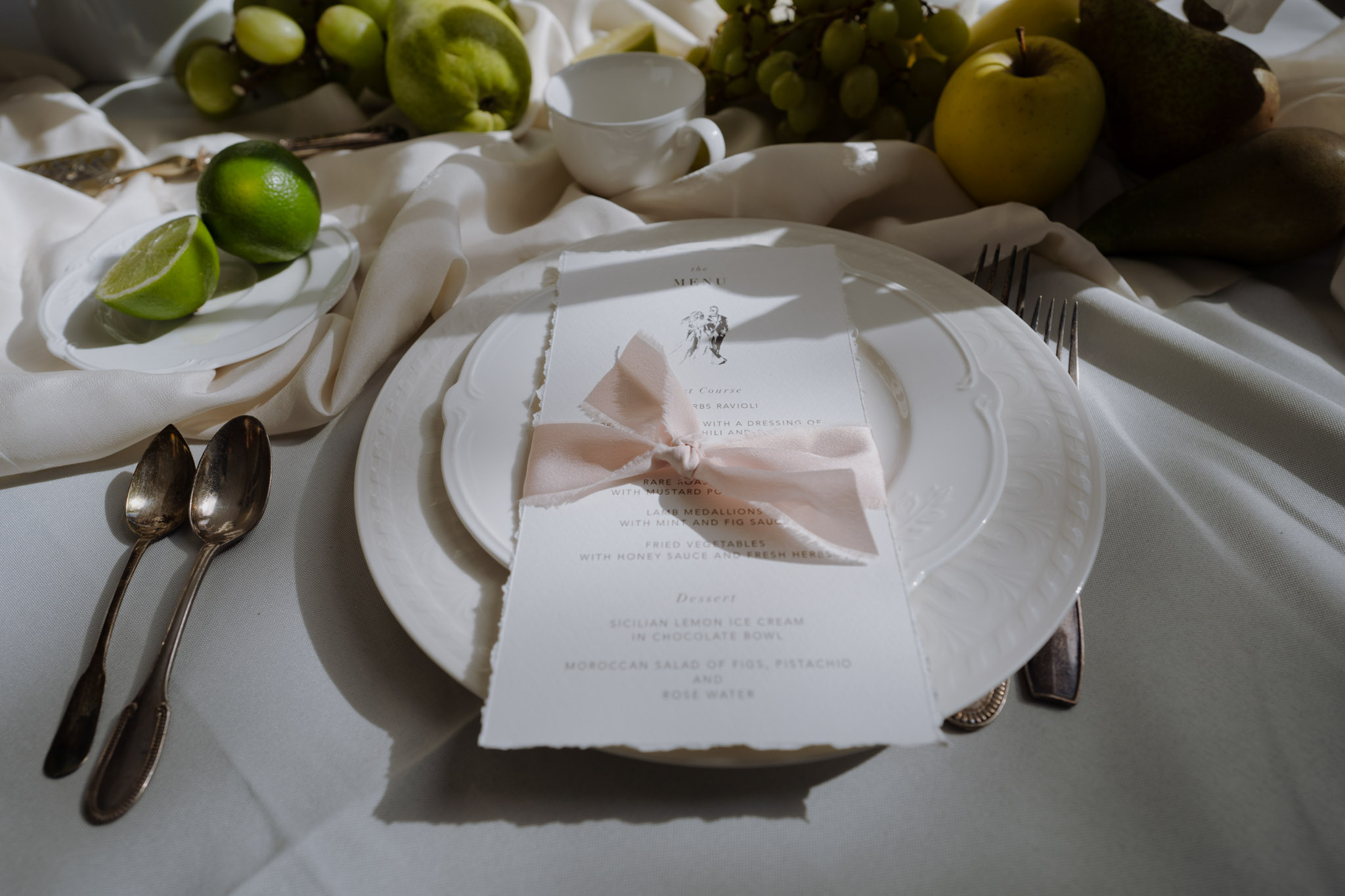 A close-up detail shot of a wedding reception place setting on a white linen tablecloth. The place setting features stacked white scallop-edged charger and dinner plates, with a deckle-edged paper menu card tied with a blush pink silk ribbon bow placed on top. The menu lists courses including herb ravioli, rare roast beef with mustard, lamb medallions with mint and fig sauce, fried vegetables, and desserts of Sicilian lemon ice cream in chocolate bowl and a Moroccan salad of figs, pistachio and rose water. Antique silver cutlery is arranged on either side of the plates. The background features loosely draped white linen fabric and a fruit-based table centerpiece incorporating green grapes, limes, green apples, and avocados, contributing to a fresh green and white color palette. The overall styling is classical with a modern editorial edge.