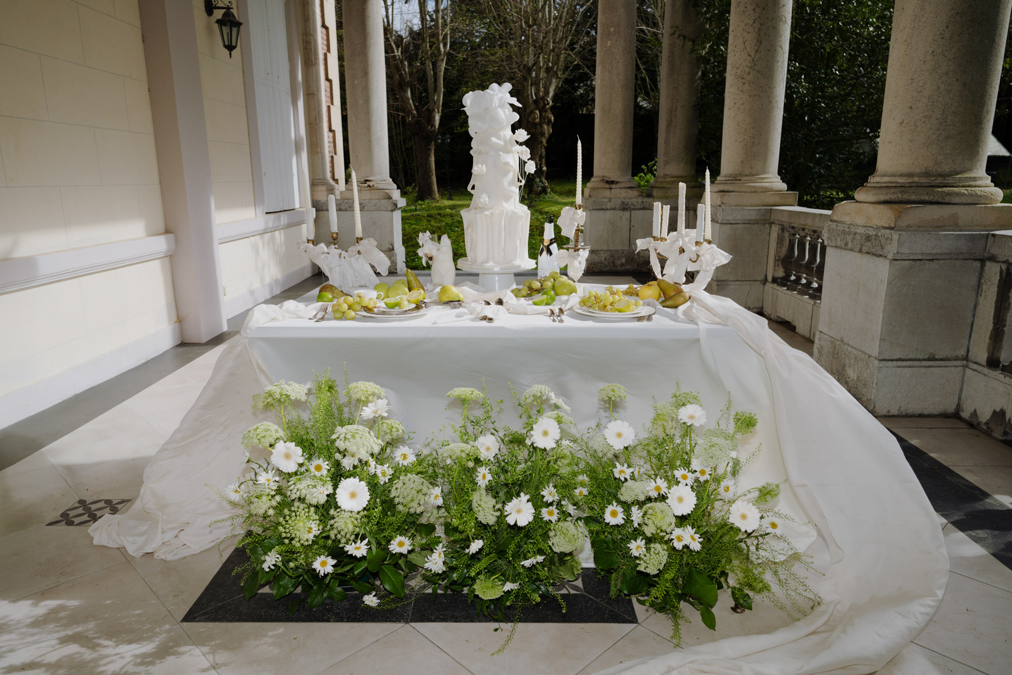 All-white ruffled cake on table with daisy and foliage installation, gold candelabras on stone portico
