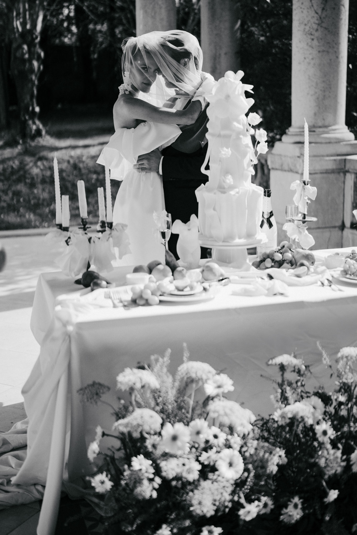 Black-and-white image of couple embracing by tiered cake on terrace with classical stone columns