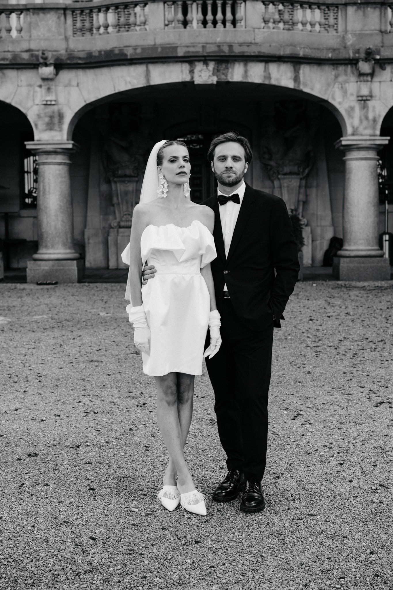 Black and white portrait of bride in short dress with cathedral veil and groom in tuxedo at stone colonnade
