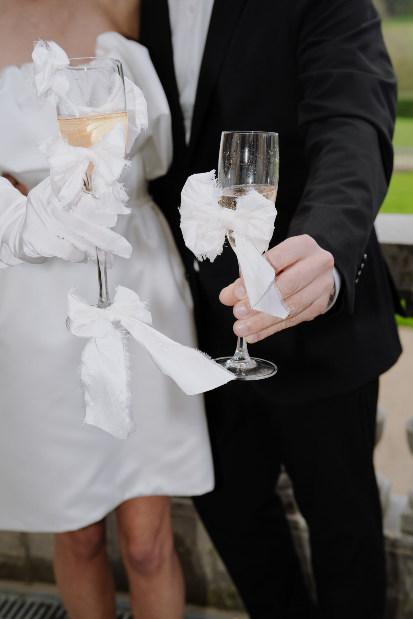 Close-up of couple holding ribbon-decorated champagne flutes, bride in white mini dress and opera gloves