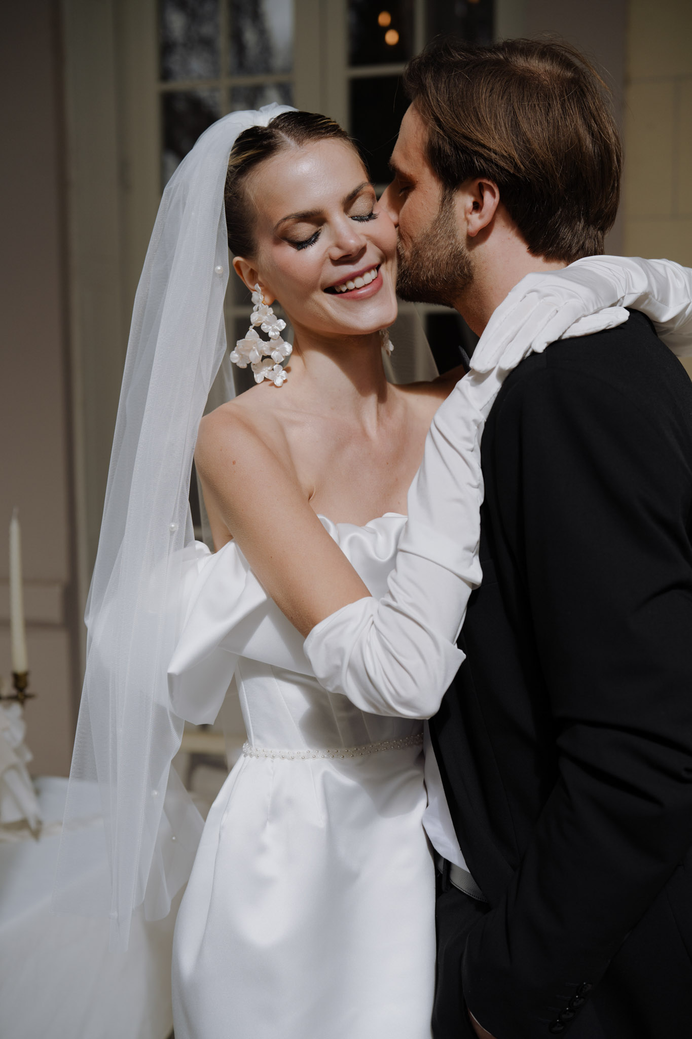 A close-up portrait of a bride and groom sharing an intimate moment indoors, with the groom kissing the bride on the cheek as she smiles with eyes closed. The bride wears a white strapless structured satin gown with a pearl-embellished waist detail, long white satin opera gloves, a cathedral-length white veil with pearl trim, and oversized white floral cluster drop earrings. The groom wears a black tuxedo jacket. The styling is modern and classic with a fashion-forward edge. In the soft background, a candelabra with white tapers is partially visible to the left, and tall French doors or windows suggest an interior reception space with warm ambient lighting. The image is shot at a medium-close portrait framing with a shallow depth of field.