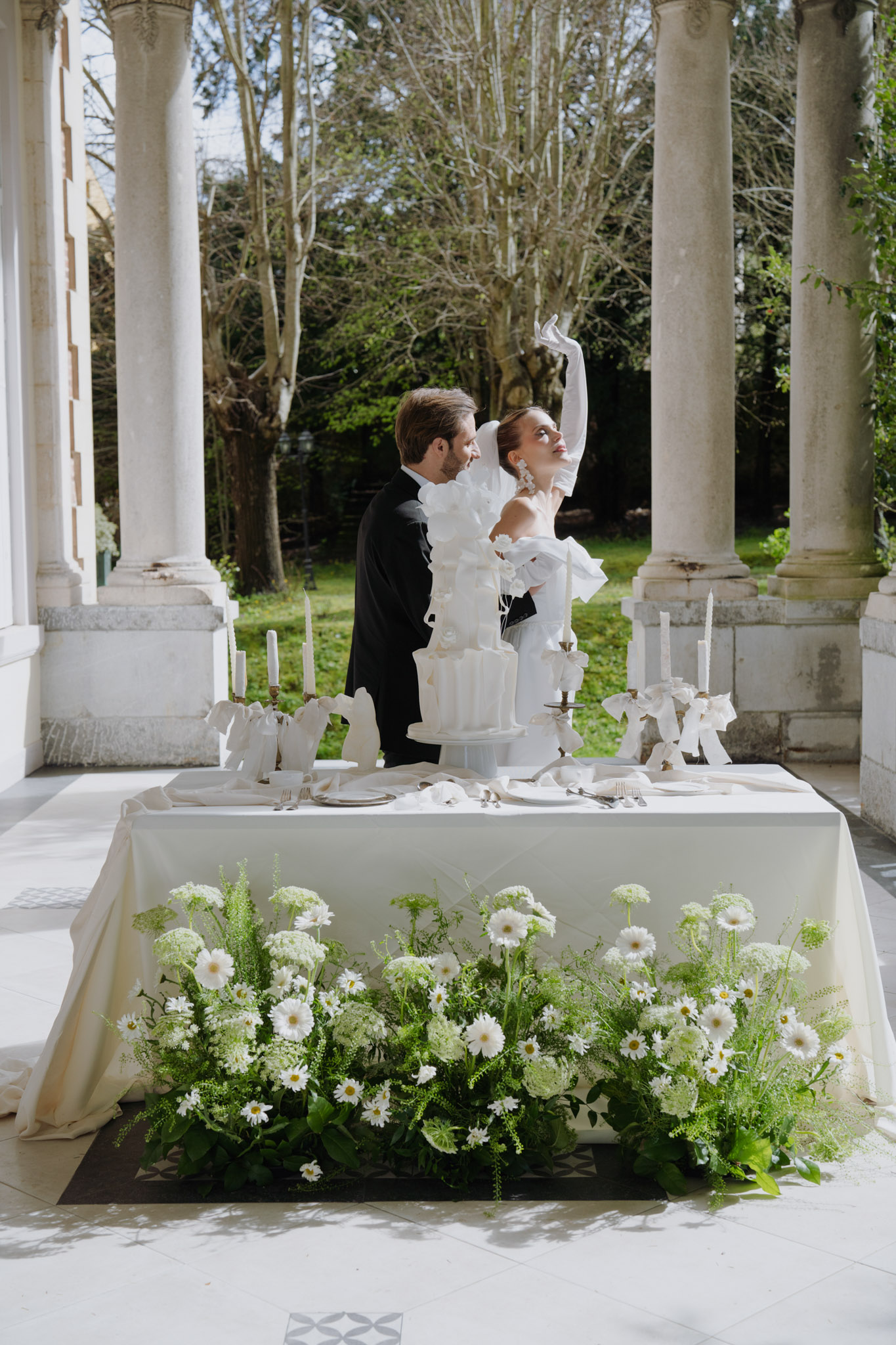 Couple at sweetheart table with white tiered cake, brass candlesticks, and white daisy ground installation on terrace