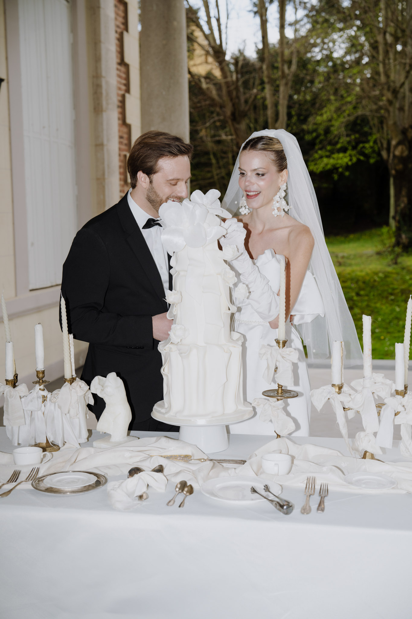 Couple behind sculpted bow wedding cake with brass candelabras and white ceramic figurines on terrace