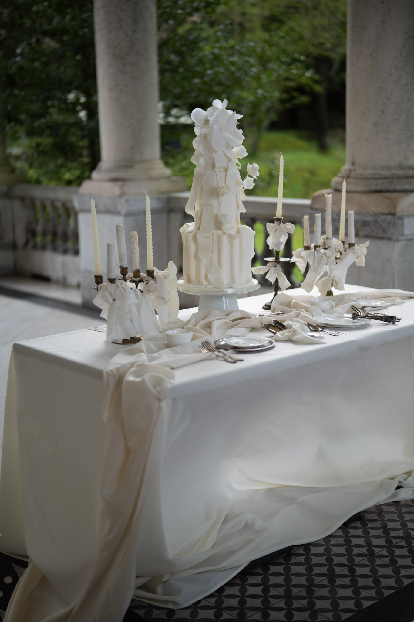 A styled wedding cake table photographed outdoors on a covered terrace or colonnade with stone columns and a balustrade. The centrepiece is a tall two-tiered white wedding cake on a white cake stand, decorated with ruffled fondant panels and large white sugar flowers. The ivory linen tablecloth drapes loosely onto a black-and-white geometric tile floor. Multiple antique brass candelabras with ivory taper candles are arranged symmetrically on either side of the cake, each adorned with loosely tied ivory ribbon or fabric bows. White porcelain plates and silver cutlery are set along the table surface. The overall styling palette is entirely ivory and white with antique brass accents, leaning toward a classic, editorial aesthetic. Medium-distance detail shot taken from a slight angle.