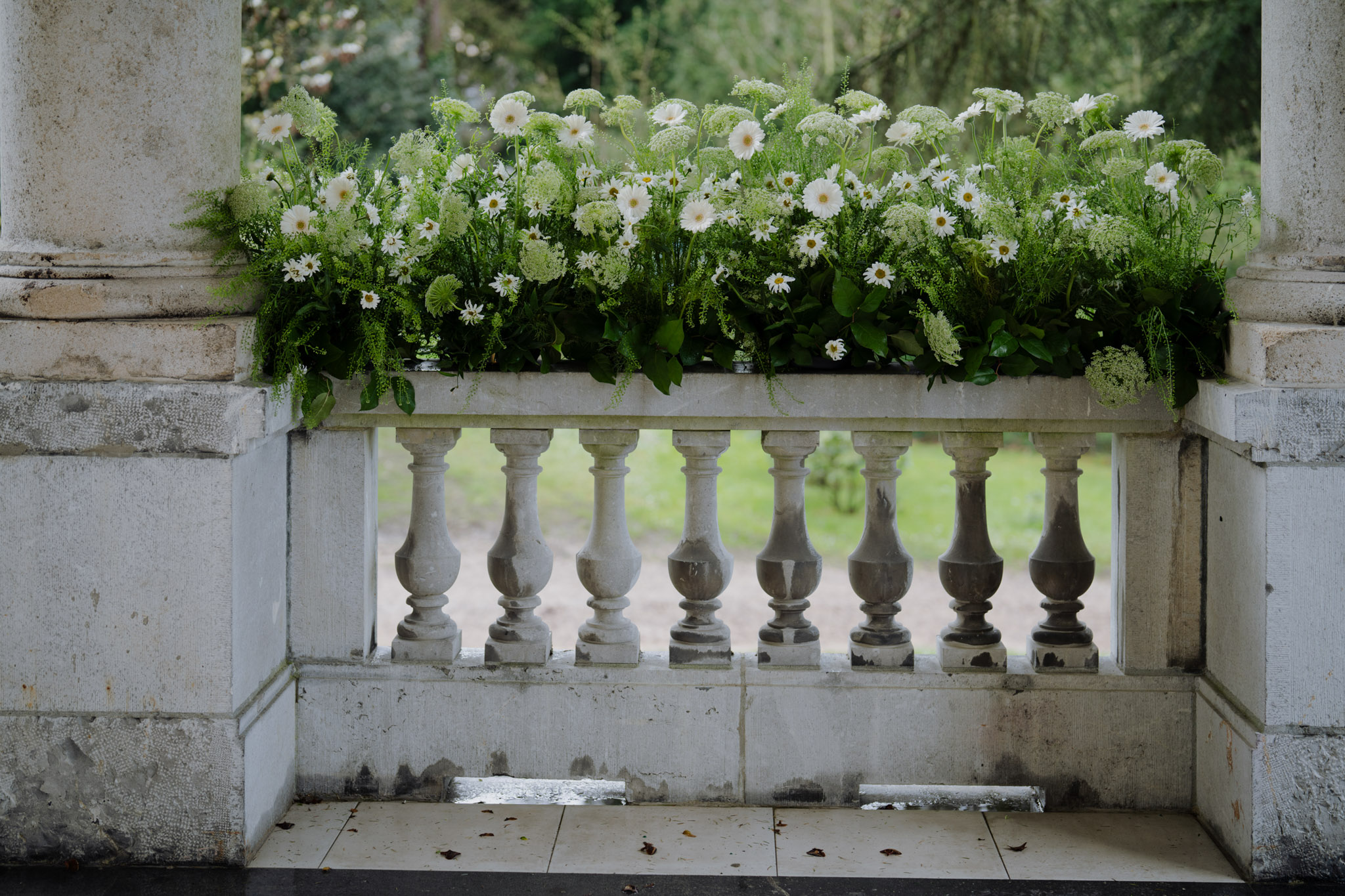 A close-up detail shot of a floral arrangement placed on top of a stone balustrade at what appears to be a château or manor house terrace. The arrangement is a lush, garden-style display composed of white gerbera daisies, white queen anne's lace, green amaranthus, ferns, and dense foliage, creating an overflowing, wildflower-inspired look in a white and green palette. The stone balustrade features turned balusters in a classical French architectural style, framed by two pilasters on either side. No people are visible; this is purely a decor and venue detail shot.