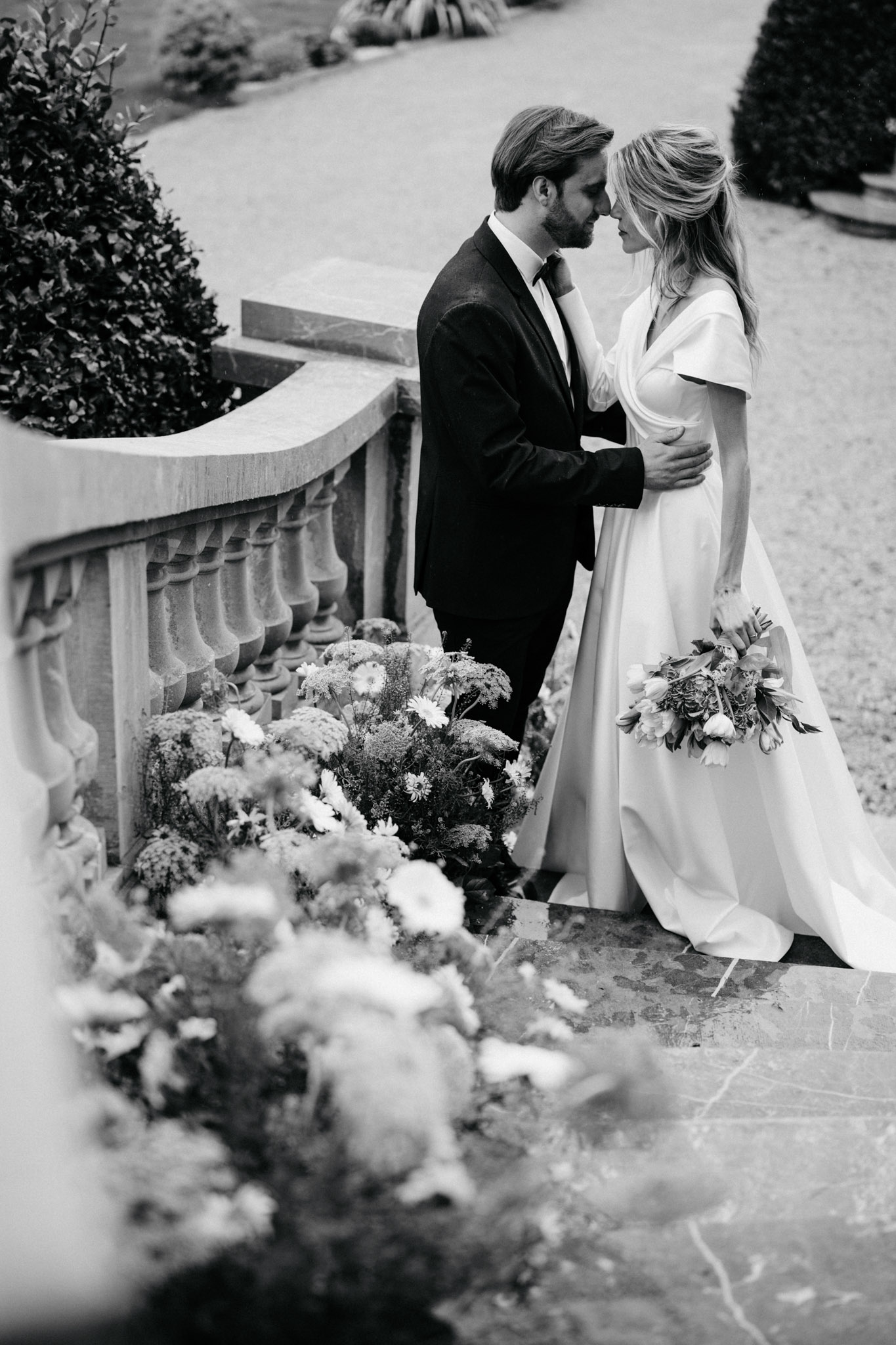 Black and white bride and groom foreheads touching on stone balustrade with garden behind