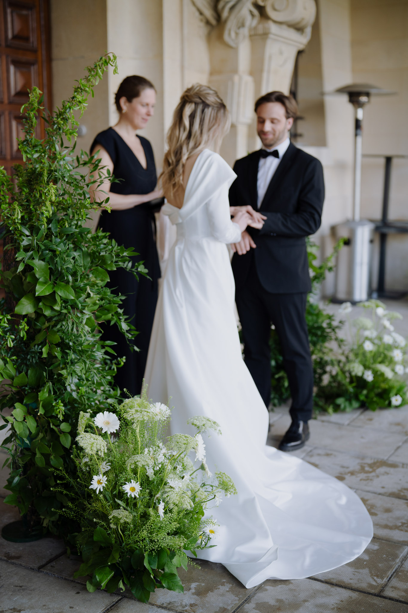 Ring exchange under stone colonnade with trailing greenery arch and white wildflower ground arrangement