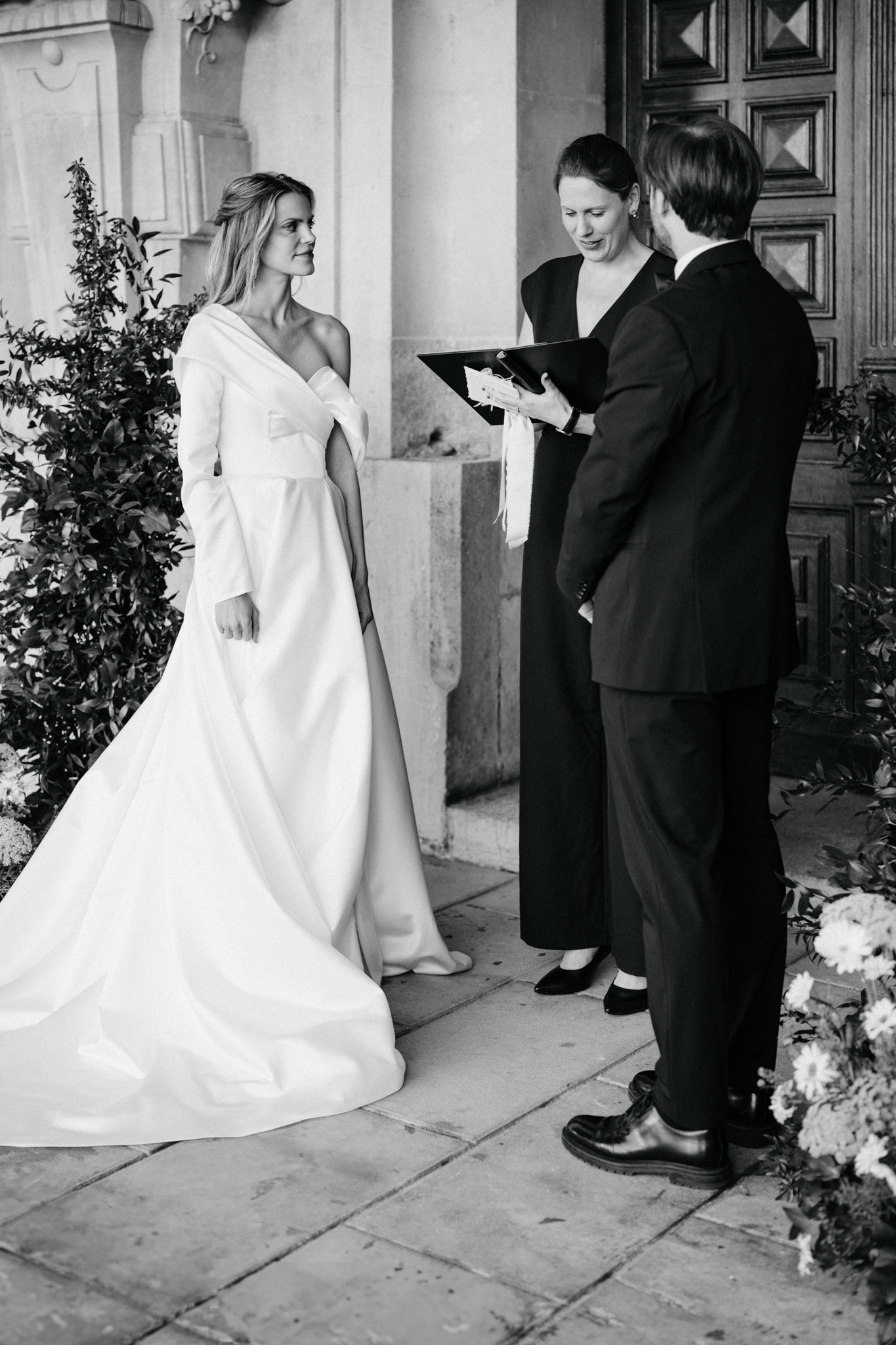 This black-and-white image captures an outdoor ceremony moment in front of a large ornate wooden door flanked by stone columns, suggesting a château or classical French estate entrance. The bride wears a structured, minimalist long-sleeve gown with an asymmetric off-one-shoulder neckline and a full skirt with a train, styled in a clean modern silhouette. The groom, in a dark suit, stands facing away from the camera alongside a female officiant dressed in dark clothing who holds a black ceremony folder with white ribbon ties. Lush foliage and a cluster of full blooms — likely hydrangeas and dahlias — are visible at the edges of the frame, and the bride looks toward the groom with a calm, direct expression. The image is a medium portrait shot with strong contrast between the bright white gown and the darker architectural background.