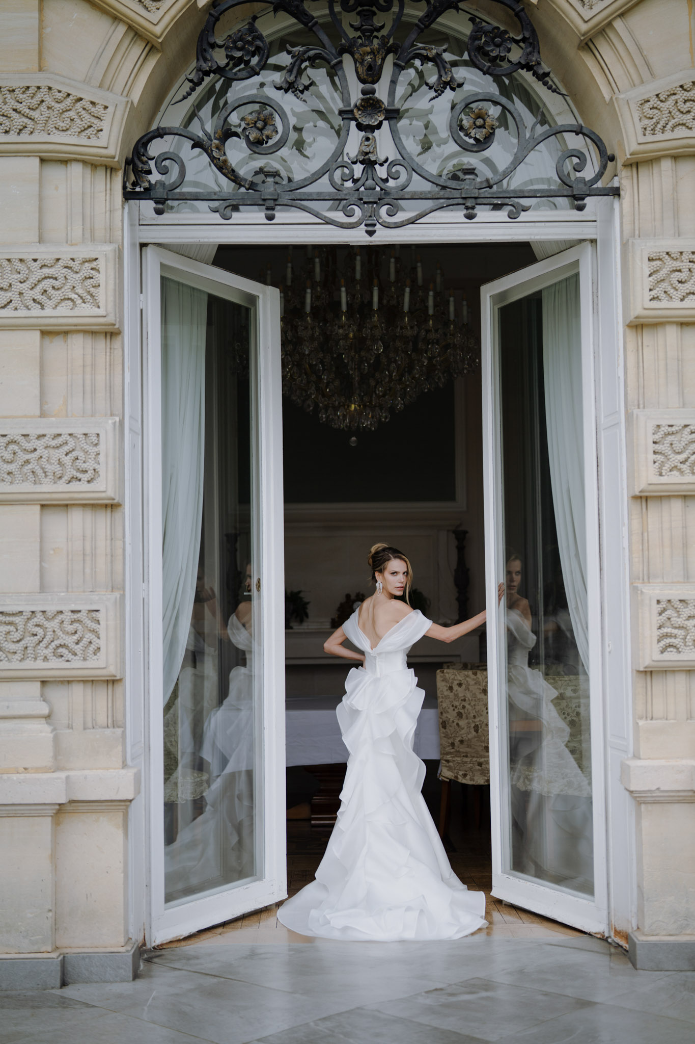 A bridal portrait of a single bride standing in the open double French doors of a château or manor house, looking back over her shoulder toward the camera. She wears a white off-shoulder structured gown with ruffled, tiered skirt panels and a floor-length train, styled with an upswept bun and statement drop earrings. The ornate carved stone doorframe is topped with an elaborate black wrought-iron fanlight in a scrollwork pattern, and a large crystal chandelier is visible hanging in the darkened interior room behind her, along with sheer white curtains and a dining table with upholstered chairs. The composition is a full-length portrait shot from outside, with the bride centered in the doorway threshold, her reflection visible in the glass door panels on either side. Potential venue feature image.