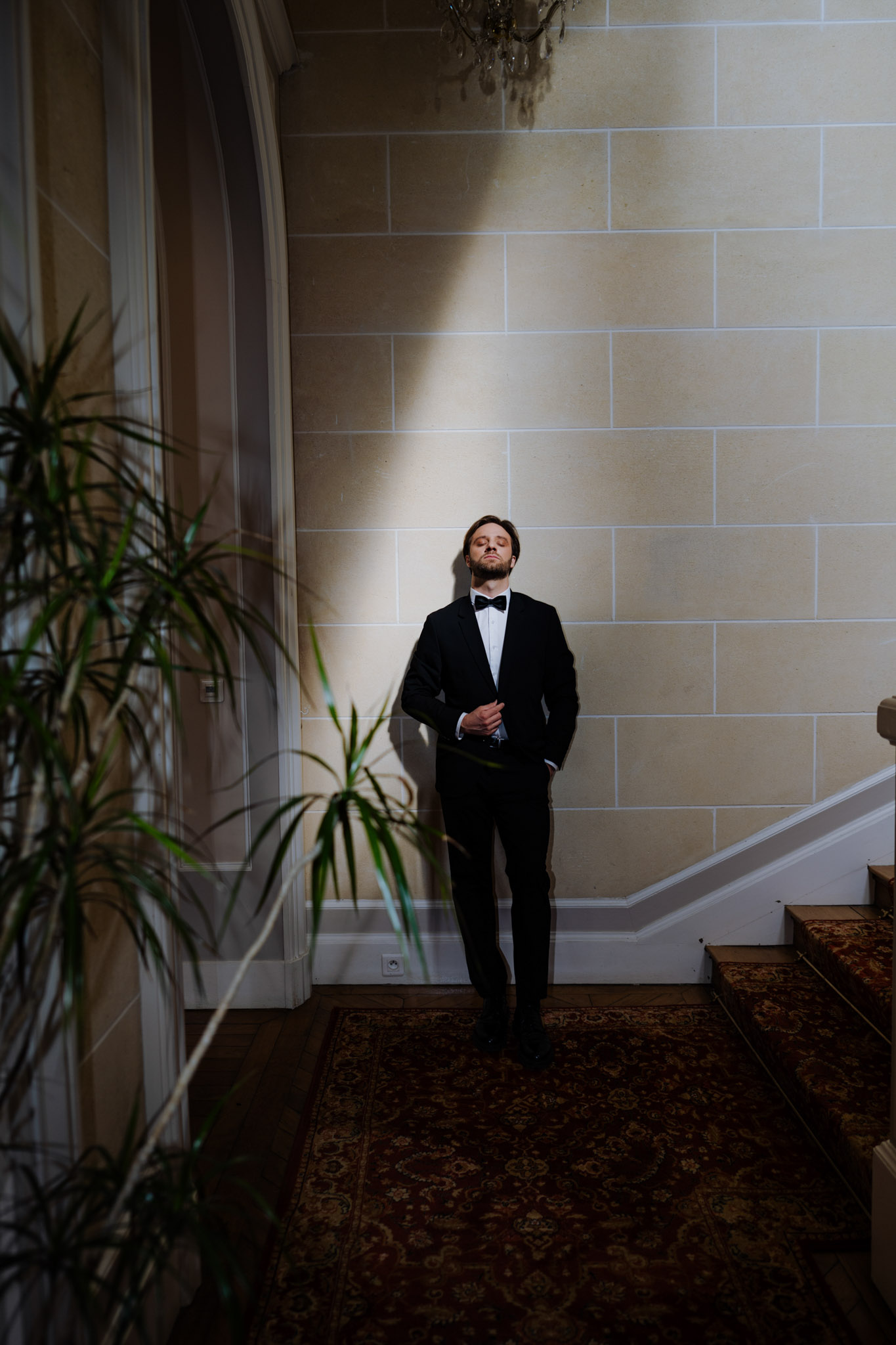 Groom in black tuxedo leaning against stone wall in chateau entrance hall with dramatic window light