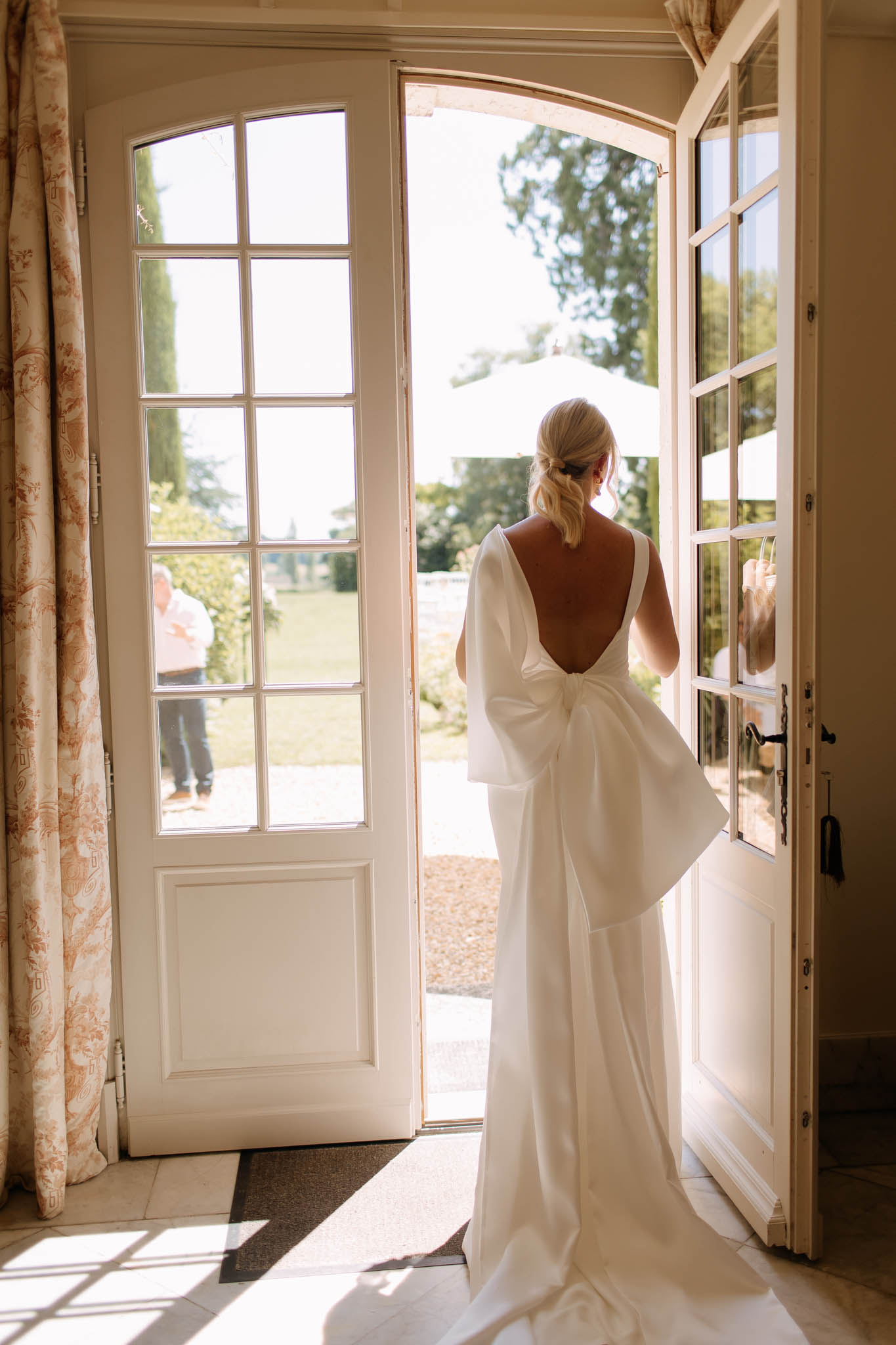 Bride photographed from behind in open-back gown with bow detail standing in doorway of French chateau