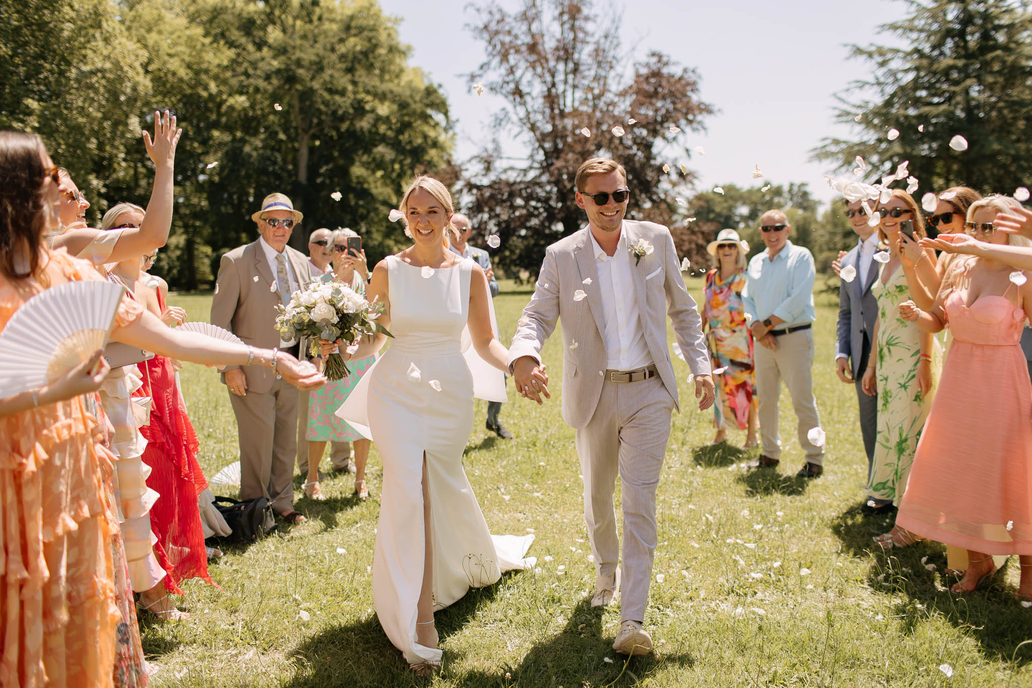 The bride and groom walk hand-in-hand through a confetti exit on the grounds of an outdoor venue, surrounded by approximately 20 guests lined up on either side throwing white flower petals. The ceremony has taken place outdoors on a lawn in bright summer sunlight, with a classic, modern aesthetic. The bride wears a sleeveless, fitted ivory satin gown with a front slit and train, carrying a bouquet of white blooms and eucalyptus; the groom wears a light sand-colored suit with a white shirt, no tie, and a white boutonniere. Guests are dressed in colorful summer attire including coral, peach, and floral-print dresses, with some holding white hand fans, suggesting a warm-weather wedding. The shot is a medium-wide portrait-style image taken at eye level, capturing the couple in the foreground with guests framing them on both sides.