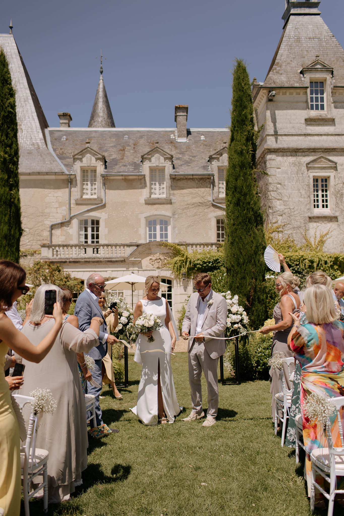 The bride and groom walk back down the aisle together following an outdoor ceremony on the lawn of a French château, with the multi-story stone building featuring pointed turrets and slate roof visible directly behind them. The bride wears a sleek white column dress with a front slit and carries a bouquet of white flowers including what appear to be peonies, hydrangeas, and eucalyptus, while the groom wears a light beige linen suit with sunglasses and white sneakers. Guests seated on white chairs line both sides of the grass aisle, with small clusters of baby's breath tied to the chair ends as aisle decor; tall black iron stands topped with white floral arrangements mark the ceremony space. The overall styling is relaxed and warm-weather classic, with guests dressed in colorful summer attire and some holding white paper fans, shot as a wide portrait-oriented image capturing both the couple and the château façade in full.