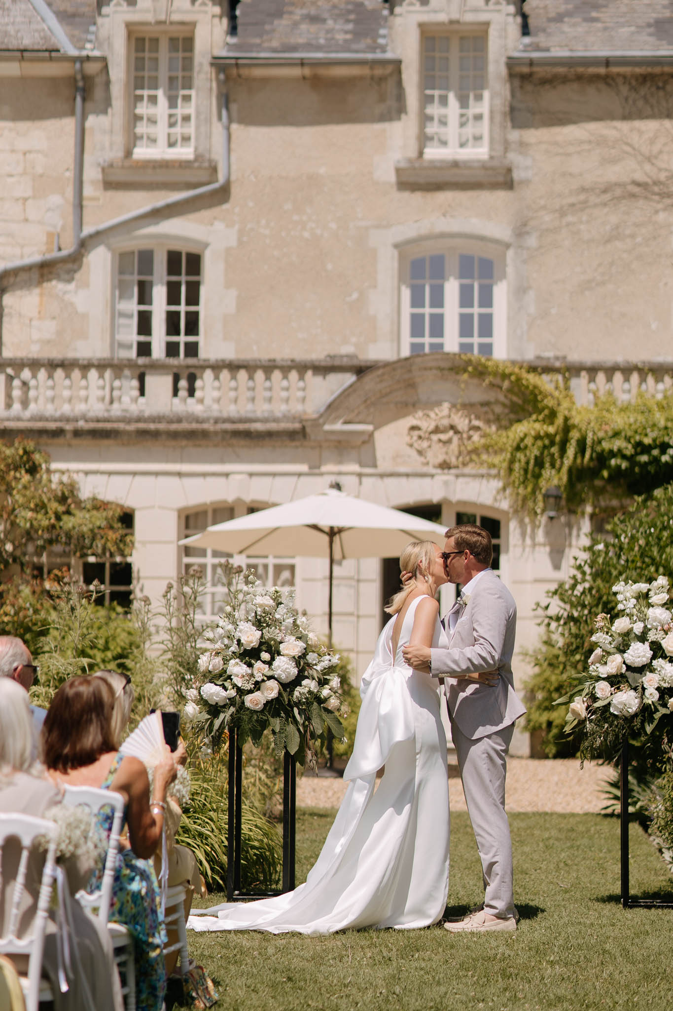 The bride and groom share their first kiss during an outdoor ceremony on the lawn of a French château. The bride wears a white sleeveless gown with a deep V-back and a long train, while the groom is dressed in a light grey suit with a pale blue tie and sunglasses. The ceremony altar is flanked by two tall black pedestal arrangements featuring white garden roses, peonies, and lush greenery. Seated guests, visible in the foreground, watch from white chairs arranged in rows, with one guest holding up a phone to photograph the moment. The backdrop is the limestone façade of a classic French château with ornate balustrades and tall white-framed windows. Wide shot capturing the full ceremony setup and venue context. Potential venue feature image.