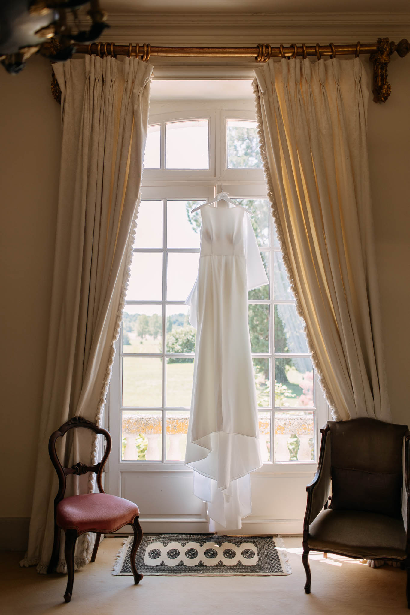 Minimal ivory gown hangs before multi-pane window with gilt rod and antique chairs in chateau room