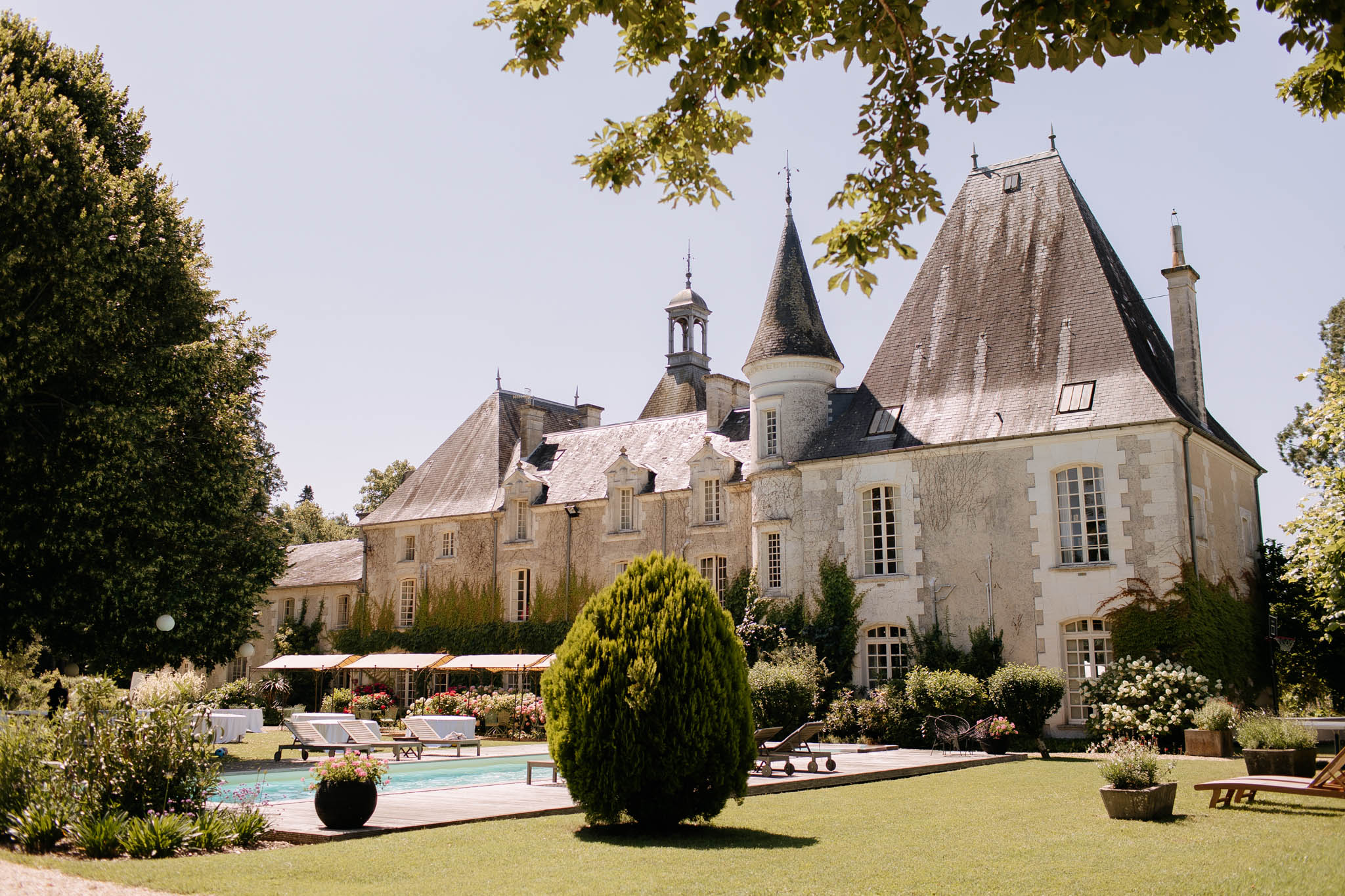 An outdoor wide shot of a French château viewed from its garden grounds, featuring cream-colored stone architecture with dark slate pointed turret roofs, dormer windows, and a central belvedere. The foreground includes a rectangular outdoor swimming pool with a wooden deck surround, sun loungers, a pergola structure draped with flowering plants in pink and red, and a large potted topiary shrub. No people are visible in the frame. The shot appears to be taken before or between wedding events, showcasing the property's grounds including manicured lawn, flowering shrubs, and planted borders. Potential venue feature image.