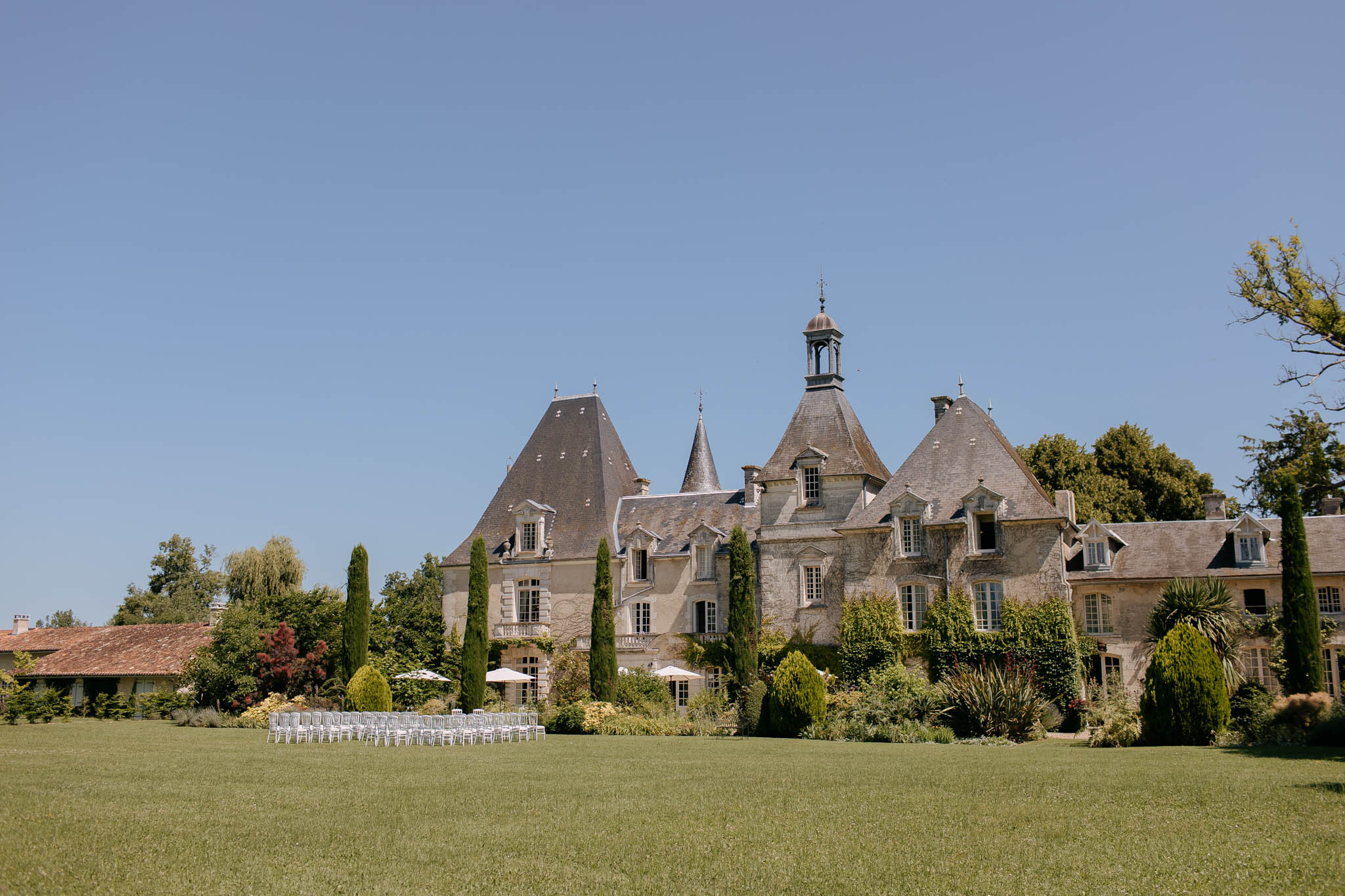 Stone chateau with pointed slate towers and cypress trees with white ceremony chairs on lawn