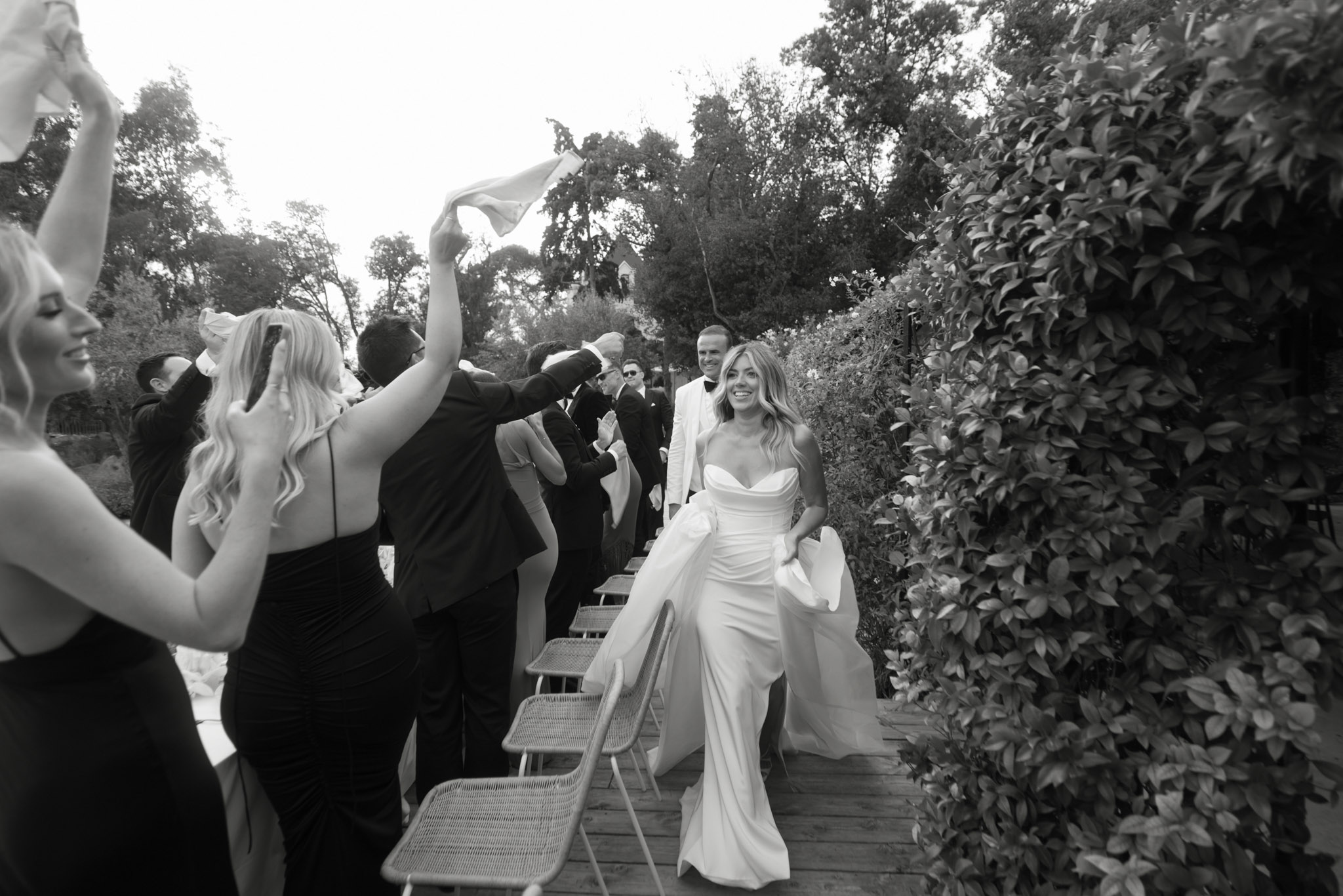 Black-and-white recessional moment with bride and groom walking through guest tunnel waving napkins outdoors
