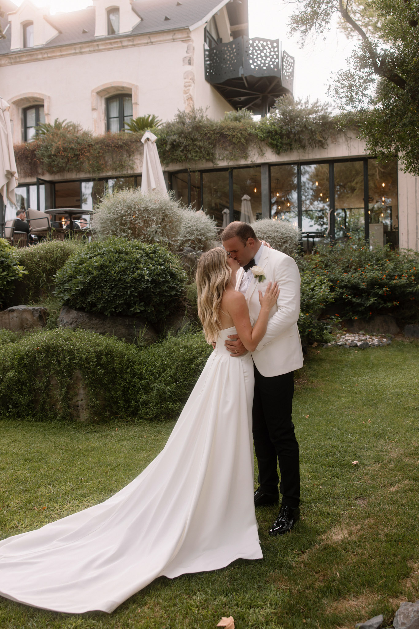 The bride and groom share a kiss during an outdoor couple portrait on a manicured lawn in front of a French stone manor or château. The bride wears a sleeveless ivory satin gown with a long cathedral-length train, and the groom is dressed in a white dinner jacket with black trousers, black bow tie, and a white rose boutonnière. The venue building behind them features arched stone windows, a modern glass conservatory extension with warm pendant lighting visible inside, a perforated metal balcony, and lush greenery cascading from upper terraces. A small group of guests can be seen seated on the terrace in the background. The styling is modern classic with a black-and-white formal palette. The shot is a full-length portrait taken from a slight distance, capturing the couple centrally framed against the venue facade.