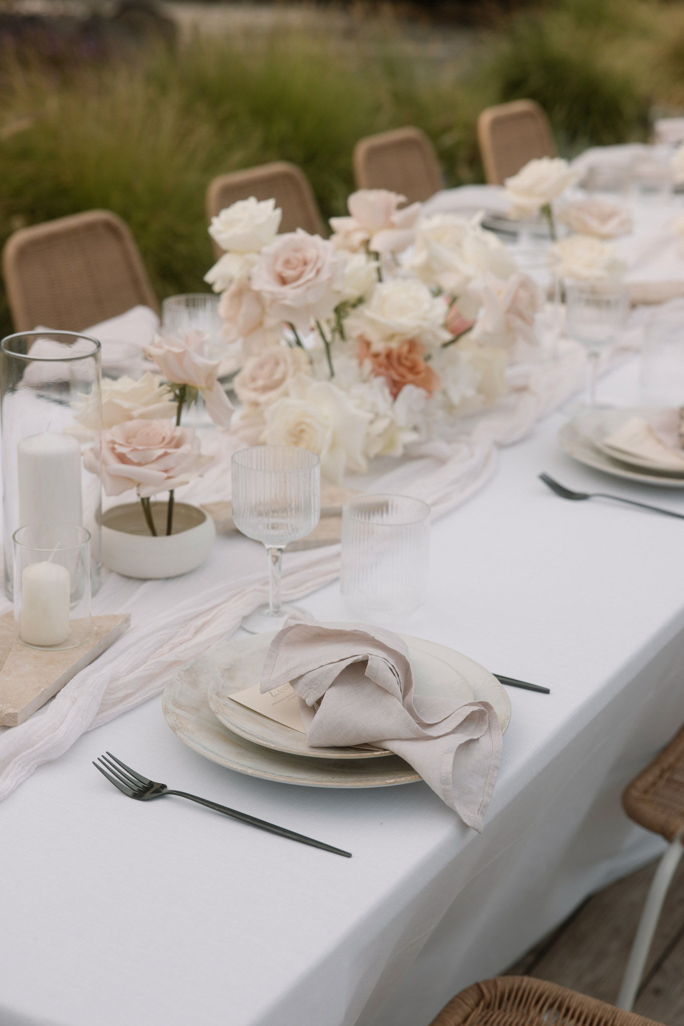 Outdoor reception table with blush roses, sand-toned plates, black cutlery, and white linen runner