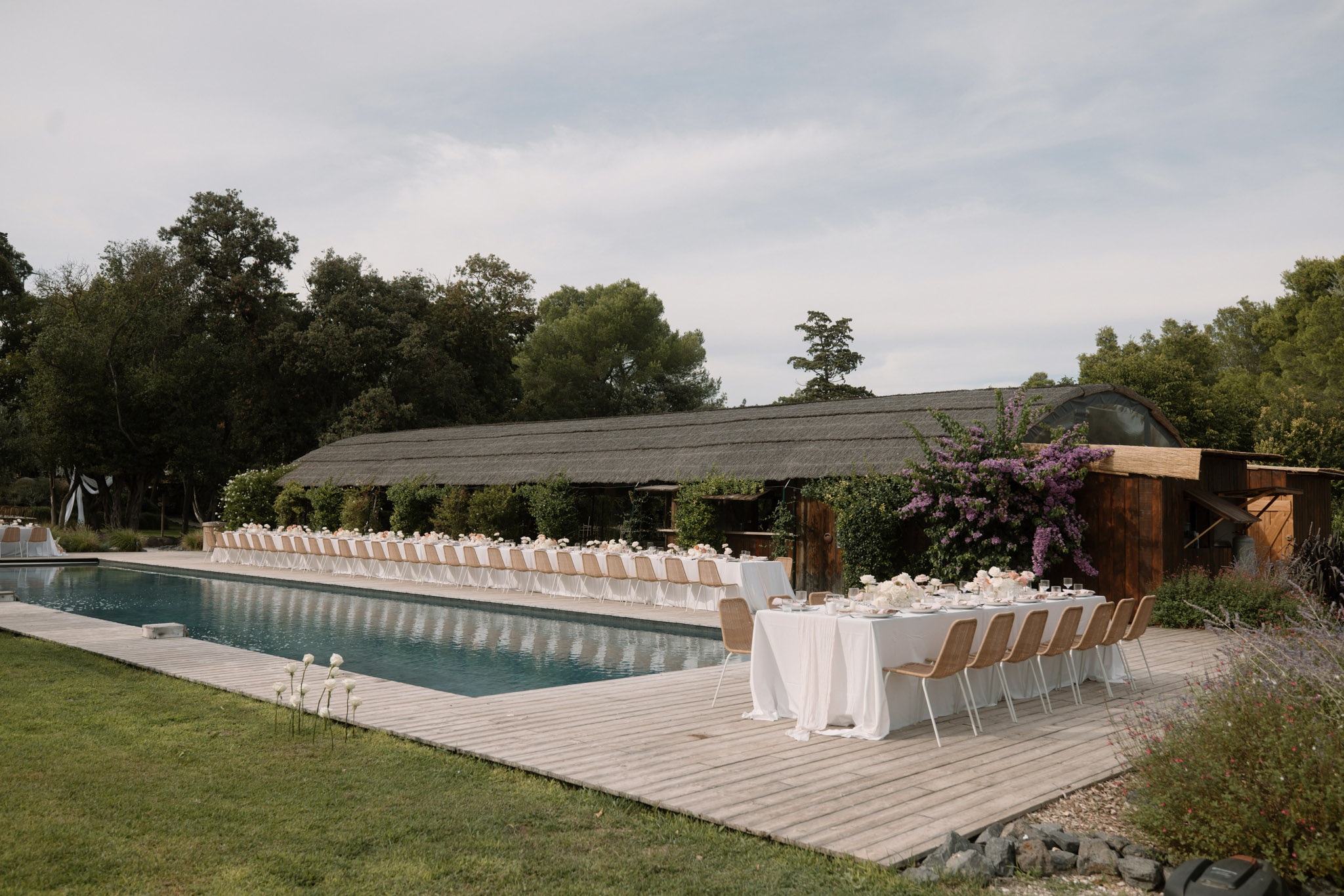 Outdoor reception tables with white linens and rattan chairs set alongside a swimming pool with rustic barn backdrop