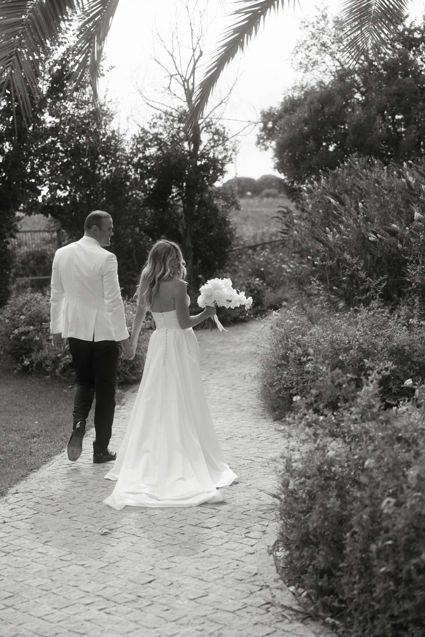 A black-and-white couple portrait taken outdoors in a garden setting, showing the bride and groom walking hand-in-hand along a cobblestone path lined with manicured shrubs and mature trees. The image is shot from behind, with the bride turning slightly to look toward the groom. The bride wears a strapless ball gown with a full skirt, button-back detailing, and a short train, and carries a round bouquet of large open blooms — likely peonies — with light ribbon wrap. The groom wears a white blazer with dark trousers and dark shoes. The high-contrast black-and-white toning gives the image deep shadows in the foliage and bright whites on the dress and jacket. Wide portrait composition with the path drawing the eye into the background.