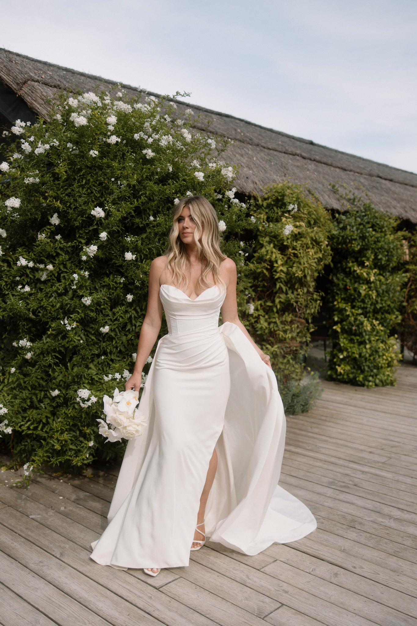 Bride in strapless ivory satin gown with thigh slit holding white rose bouquet on wooden deck
