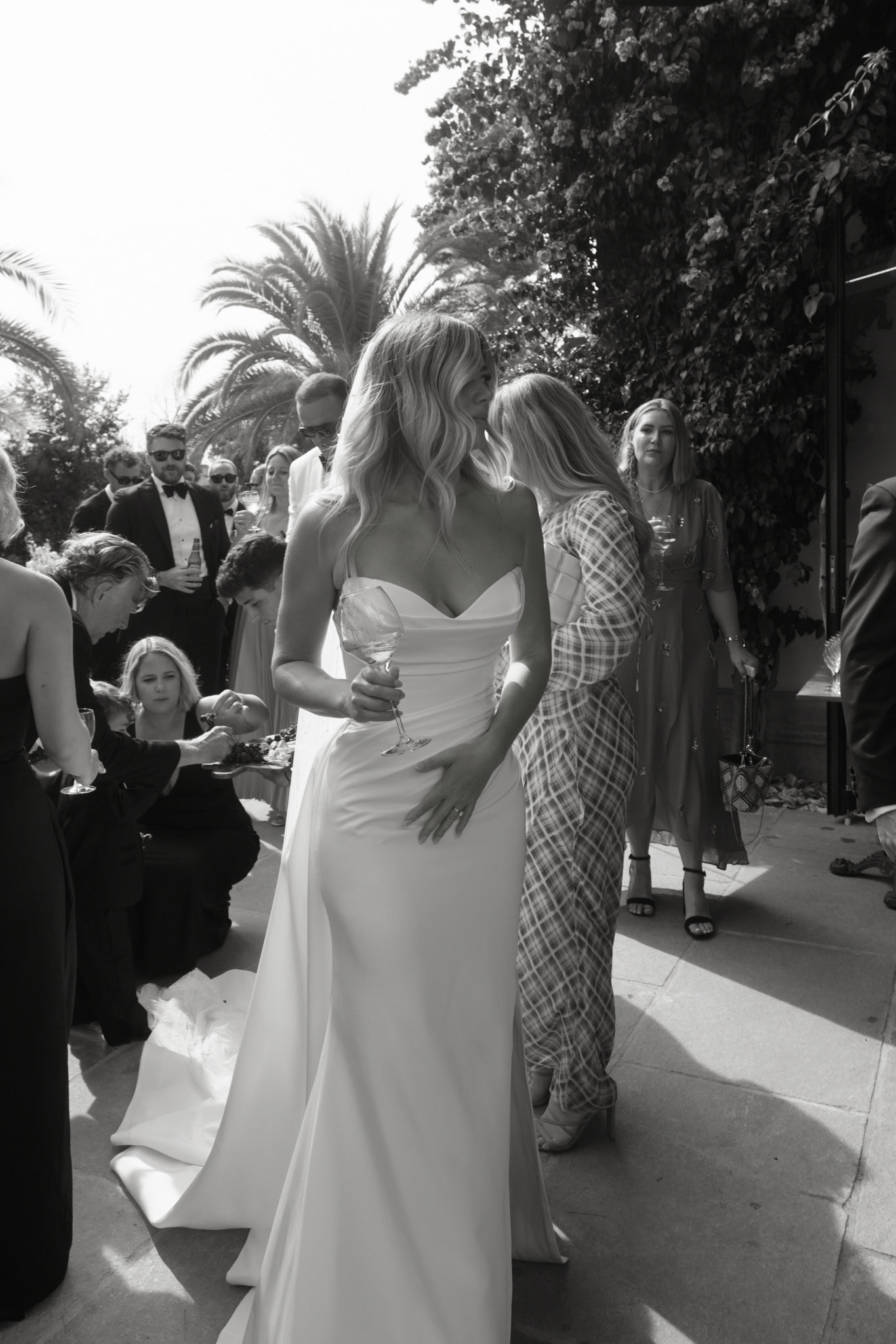Black-and-white candid of bride holding wine glass on terrace surrounded by guests in formal attire