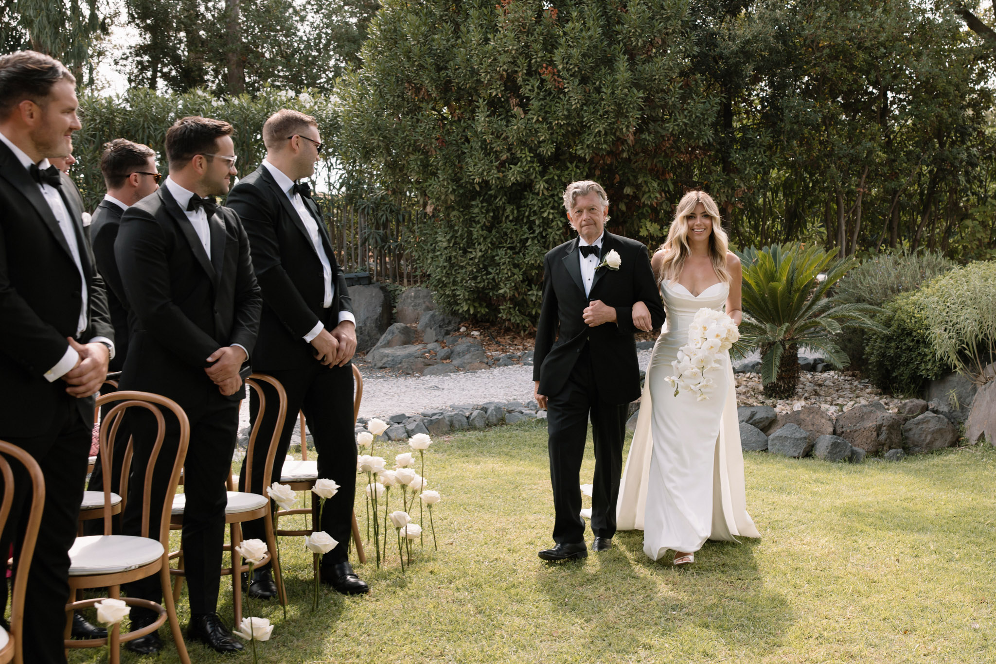 Bride walked down the aisle by her father during outdoor garden ceremony with white rose-lined path and groomsmen