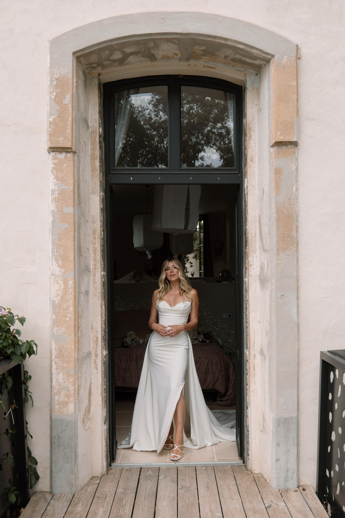 A bridal portrait of a bride standing in the open doorway of what appears to be a French château or manor house, photographed from outside. She is wearing a fitted ivory satin strapless gown with a cowl neckline, a front thigh slit, and a detachable cape or train that falls behind her. Her footwear consists of white strappy heeled sandals. She has long blonde wavy hair and is looking slightly upward with her hands clasped in front of her. The arched doorway features dark metal-framed glazing above and weathered stone surrounds with aged plaster, characteristic of a historic French property. The interior room visible behind her is dimly lit. The composition is a full-length portrait shot, centered and symmetrical within the architectural frame of the doorway.