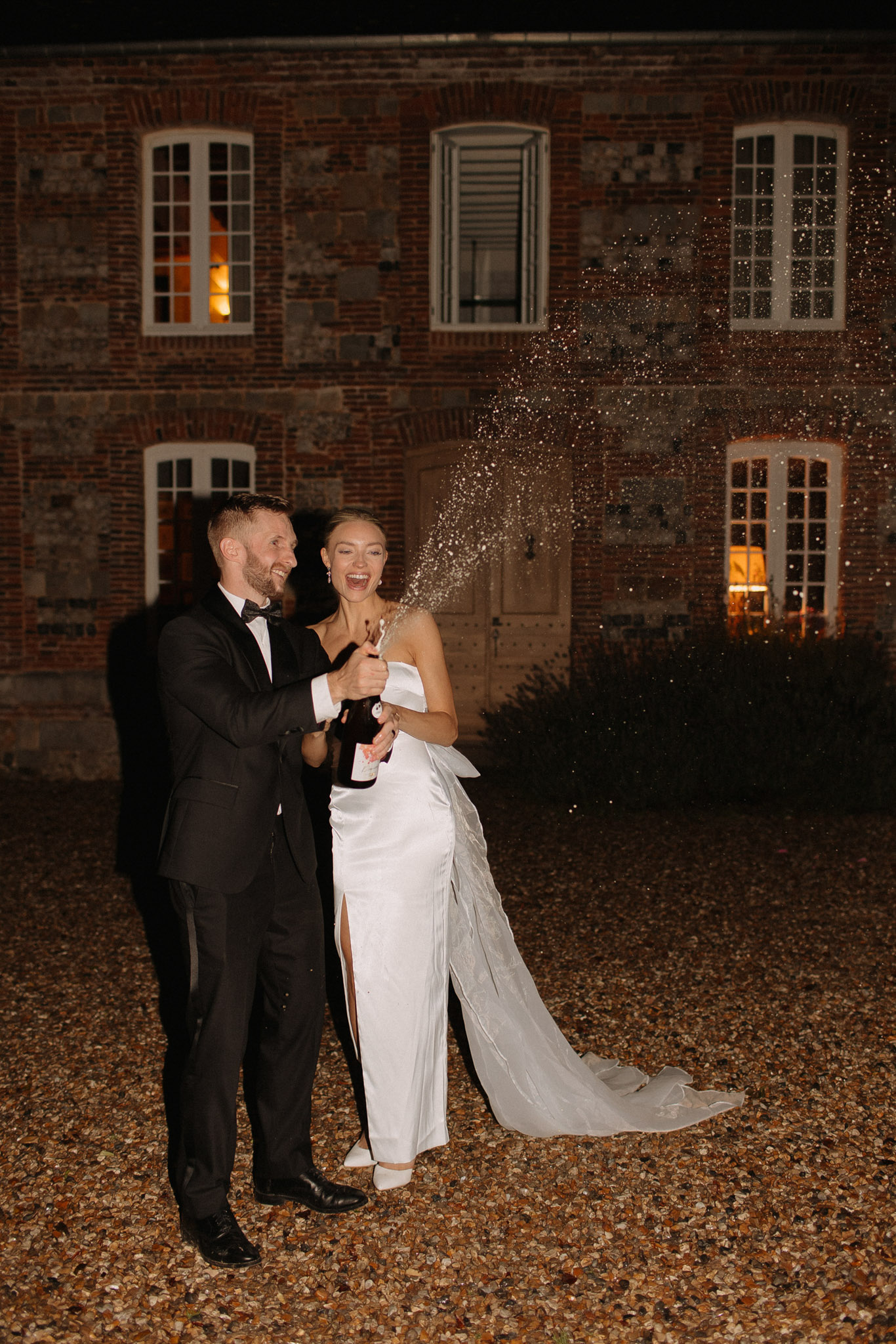 The couple is popping a champagne bottle together outdoors at night, both laughing as the champagne sprays outward. The setting is a gravel courtyard in front of a French brick and stone manor building, with warm orange light glowing from several windows. The groom wears a black tuxedo with a bow tie, and the bride wears a white satin slip-style gown with a front slit, a large bow detail at the back, and a flowing train, paired with white heels. The styling is modern and minimal, with a black-and-white color palette for the couple's attire. The image is a full-length portrait shot taken at night with flash, capturing the motion of the champagne spray.