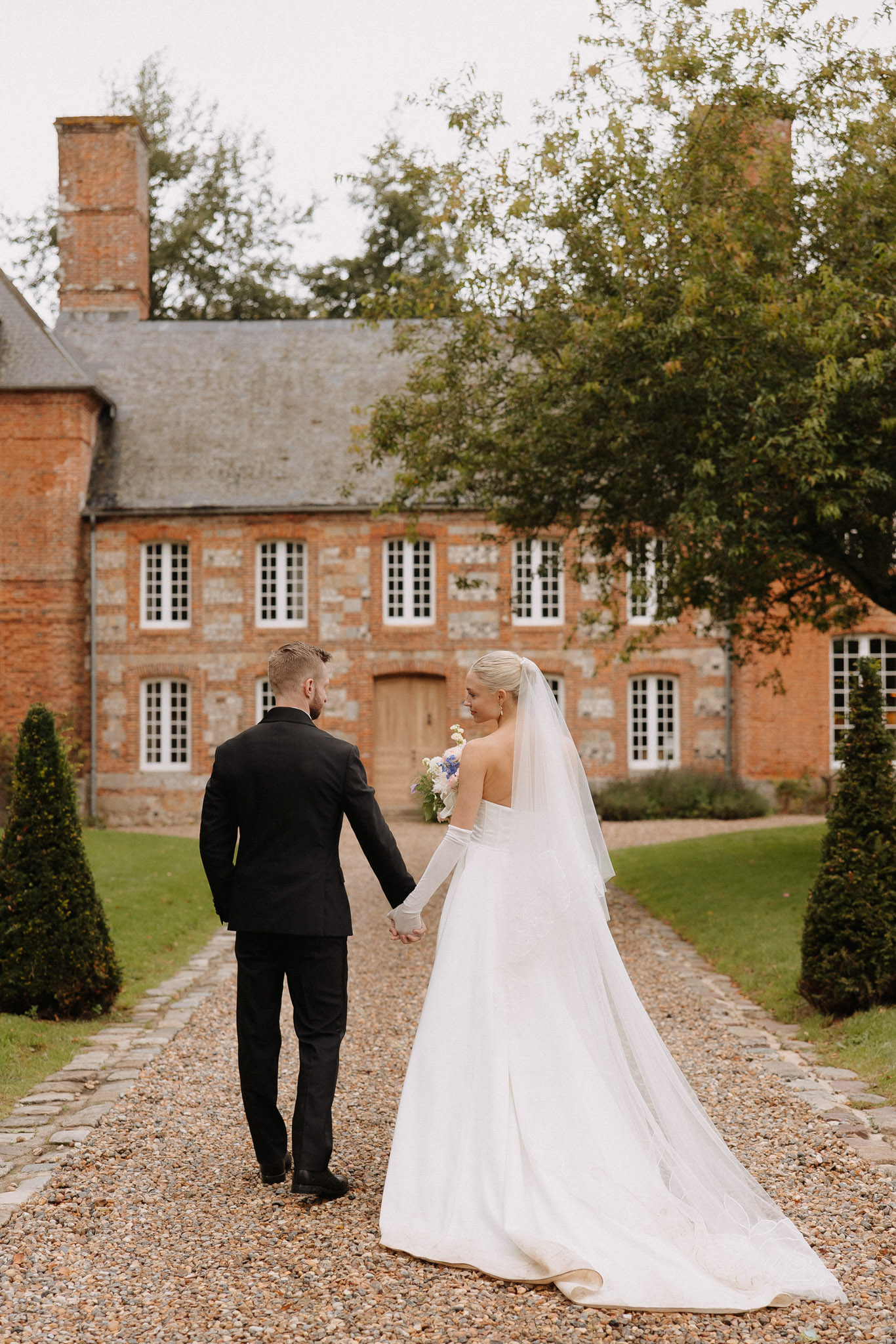 A couple portrait taken outdoors on a gravel path leading toward a two-storey red brick French manor house with white-framed windows and a slate roof. The couple is photographed from behind, holding hands and walking toward the building, with the bride turning slightly to look back over her shoulder. The groom wears a black suit, and the bride wears a white strapless ball gown with a long cathedral-length train, white opera gloves, and a simple cathedral veil; her blonde hair is styled in a sleek low updo. She carries a small bouquet featuring white and blue flowers, likely including delphiniums and white blooms. The styling is classic and minimal, with a clean white and black color palette. The composition is a full-length portrait shot from mid-distance. Potential venue feature image.