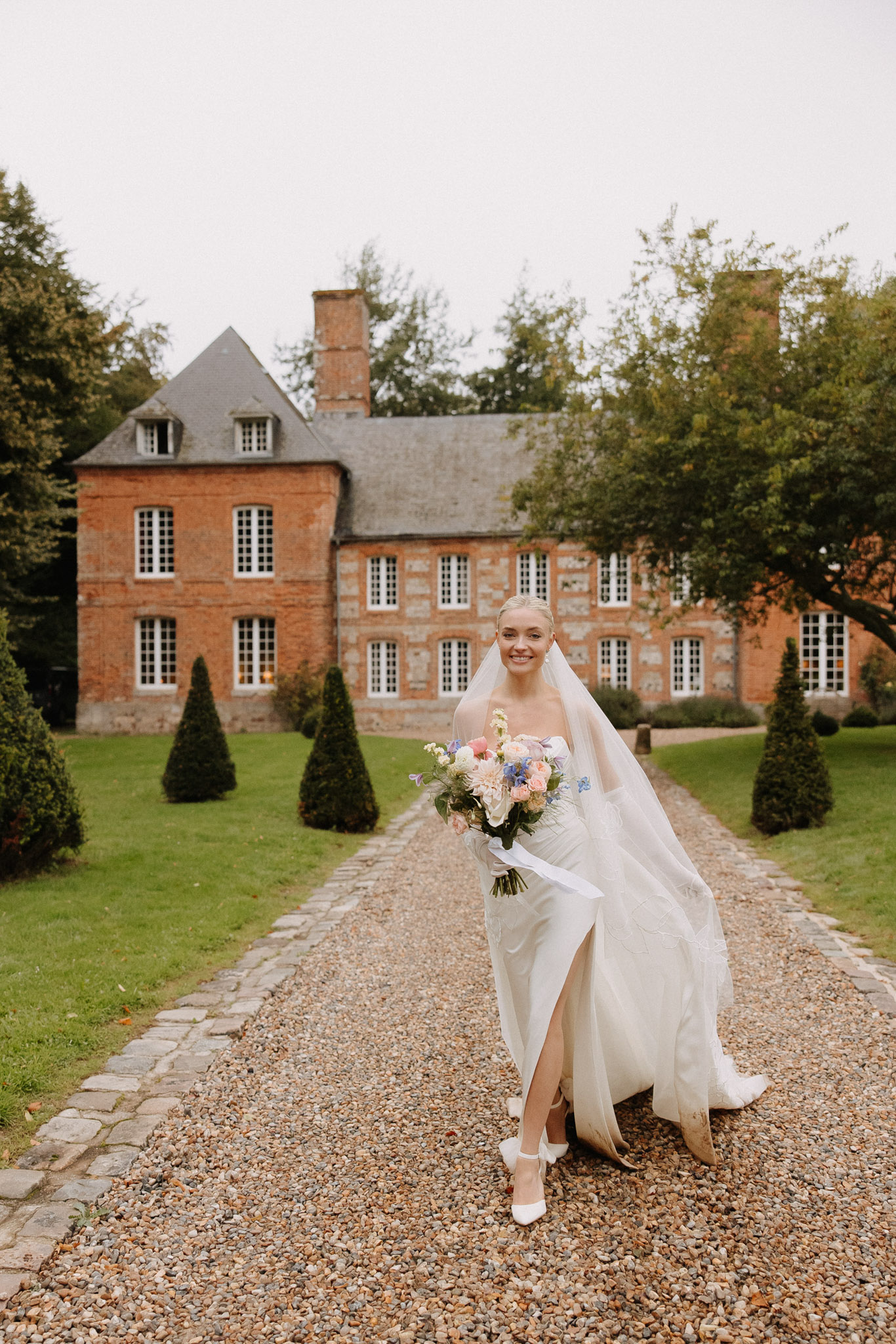 Bride walking toward camera on gravel drive of a red brick French manor holding a garden-style bouquet with blush roses