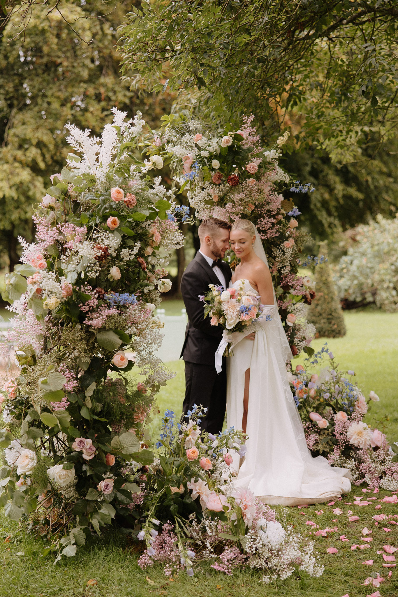Couple under circular floral arch of peach roses blue delphiniums and ivory blooms bride in opera gloves with garden bouquet