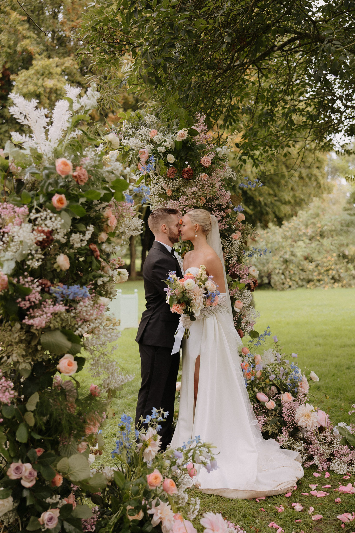 A couple shares a kiss during an outdoor ceremony portrait, standing beneath a large circular floral arch set on a manicured lawn. The groom wears a black suit with a white shirt and tie, while the bride wears a white strapless fitted gown with a front slit, a cathedral-length veil, and long white gloves. The bride carries a loose bouquet featuring peach garden roses, blue delphiniums, dahlias, and gypsophila tied with a white ribbon. The floral arch and ground-level installations are densely arranged with peach and coral garden roses, burgundy blooms, blue delphiniums, pink gypsophila, white astilbe, lavender roses, and abundant greenery, with scattered pink petals on the lawn around the base. The overall floral palette combines soft pastels with deeper jewel tones in a garden-style, romantic aesthetic. The shot is a full-length portrait framing the couple centrally within the arch.