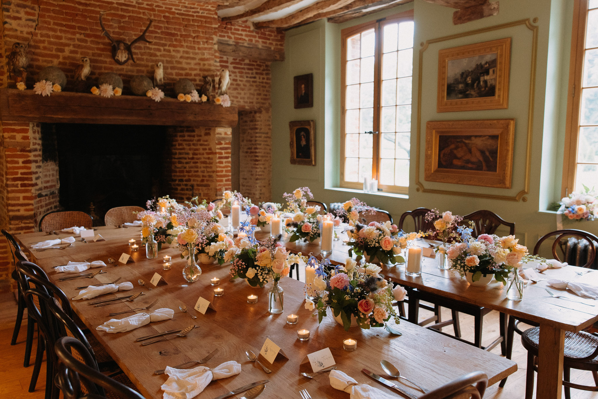 A wedding reception tablescape set inside a rustic interior room with exposed brick walls, wooden ceiling beams, and sage green painted walls hung with gilt-framed paintings. The long wooden farm table is laid with white linen napkins, silverware place settings, small place cards, and clusters of mixed floral arrangements in glass bud vases and low ceramic vessels featuring peach, coral, blush pink, and soft yellow roses alongside blue delphinium, white blooms, and baby's breath. Pillar candles on small risers and scattered tea light votives run the length of the table. The large brick fireplace in the background is decorated with pale pink and cream flower heads and taxidermy owl figures beneath a set of antlers. Dark bentwood and cane-back chairs surround the table. Wide-angle interior shot with no guests present.