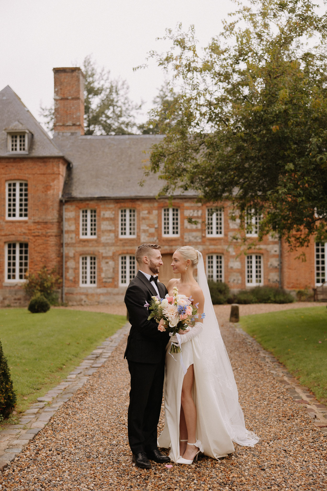 Bride with long gloves and peach dahlia bouquet and groom in tuxedo before red-brick manor with slate roof
