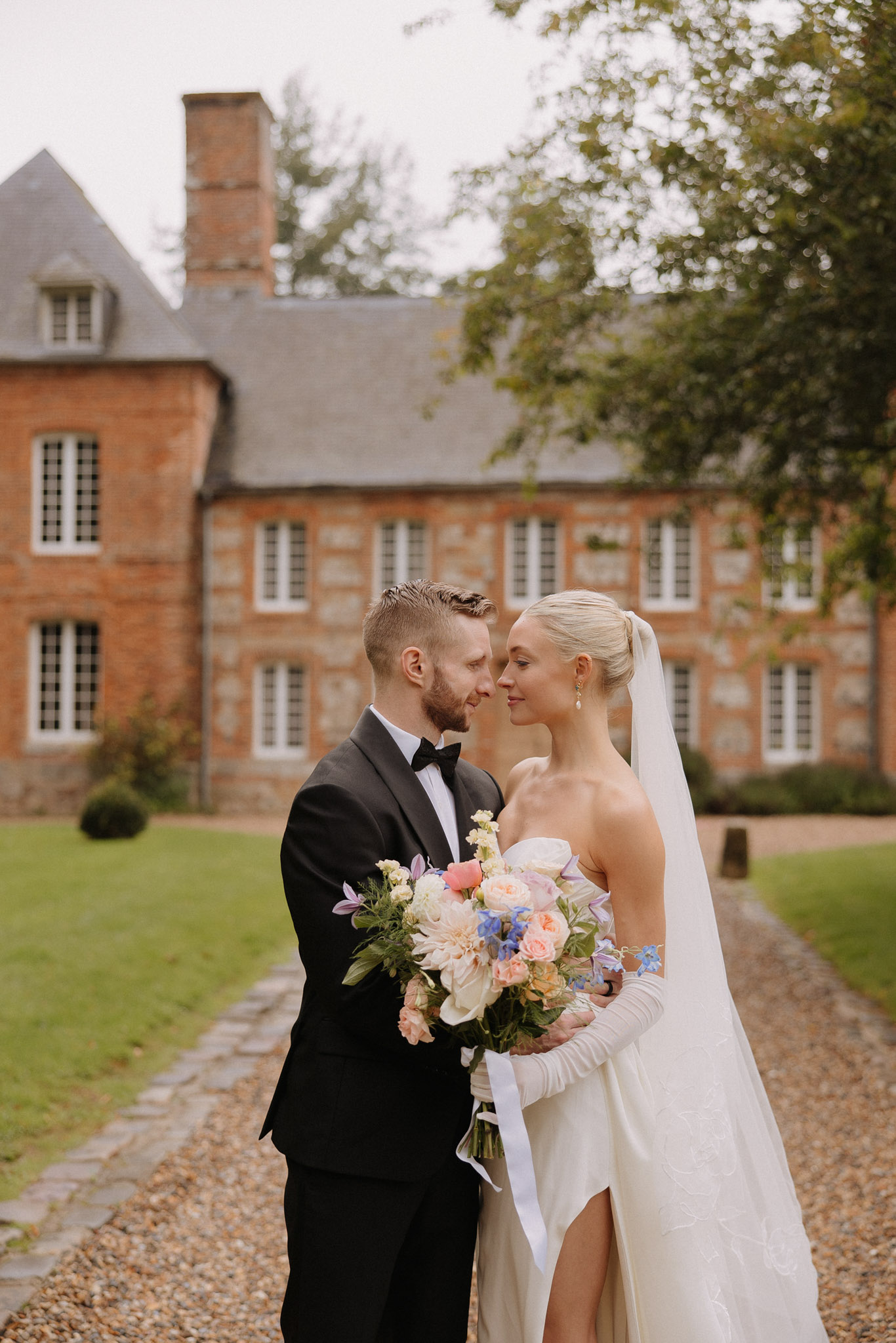 Bride in opera gloves with peach and blue bouquet and groom in tuxedo touch foreheads before manor