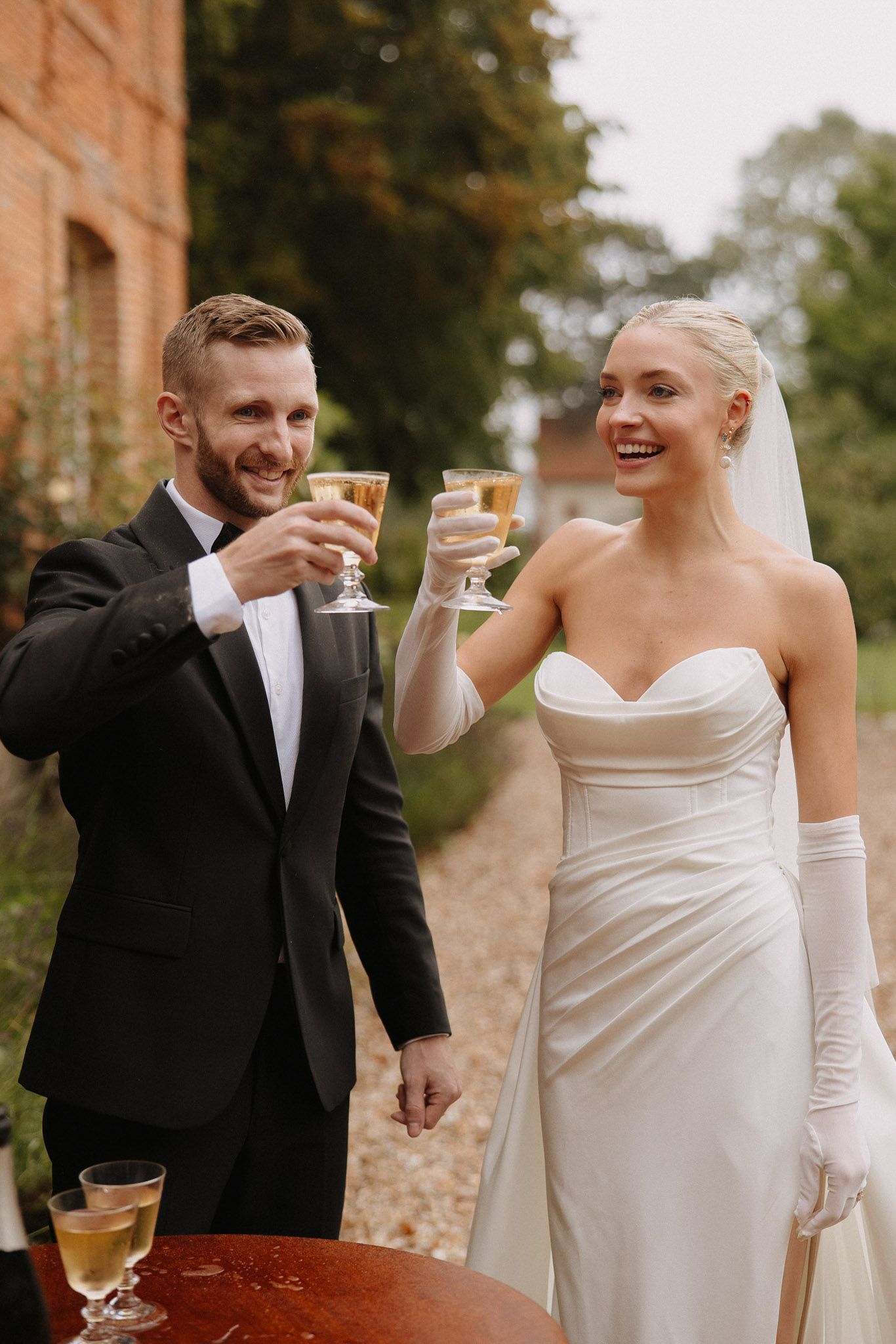 The bride and groom are toasting with champagne glasses outdoors, likely during a cocktail hour, set against the grounds of a red brick manor or château visible on the left. The groom wears a black tuxedo with a white dress shirt, while the bride wears a fitted ivory strapless satin gown with a sweetheart neckline, long white opera gloves, drop earrings, and a sheer veil with her hair pulled back. The styling is classic and formal, with a black-and-white color pairing accented by the white gloves. Additional champagne glasses are visible on a round wooden cocktail table in the foreground. This is a mid-shot portrait of the couple from the waist up.