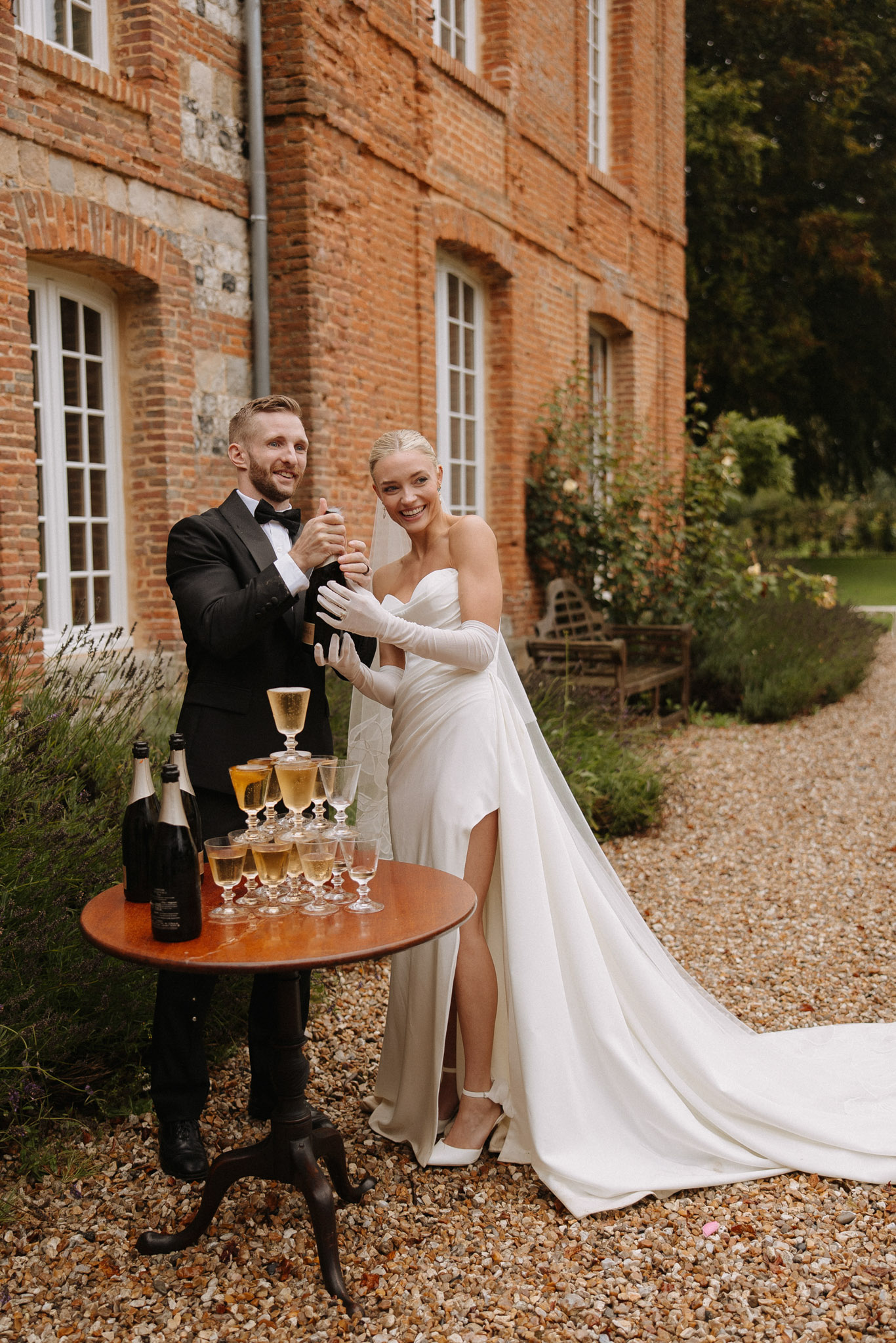 Bride in satin column gown and groom in tuxedo opening champagne beside coupe pyramid at red brick manor