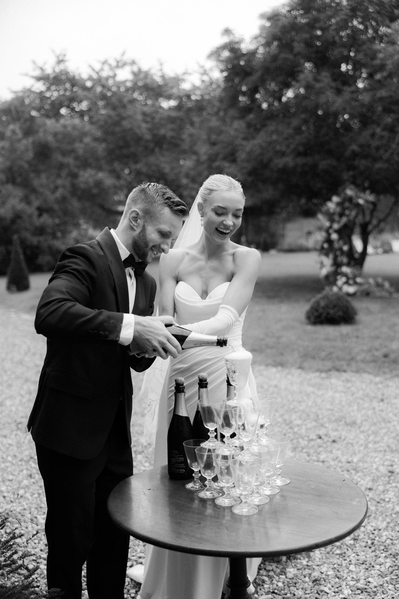 Bride in strapless gown and opera gloves with groom opening champagne over coupe tower, black and white