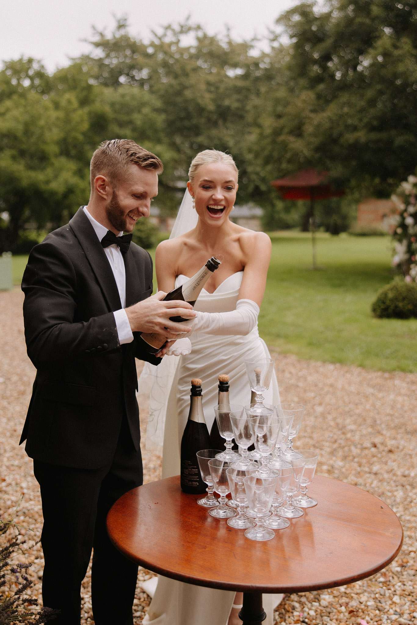 Couple opening champagne on gravel path, bride in strapless gown with white gloves beside coupe tower