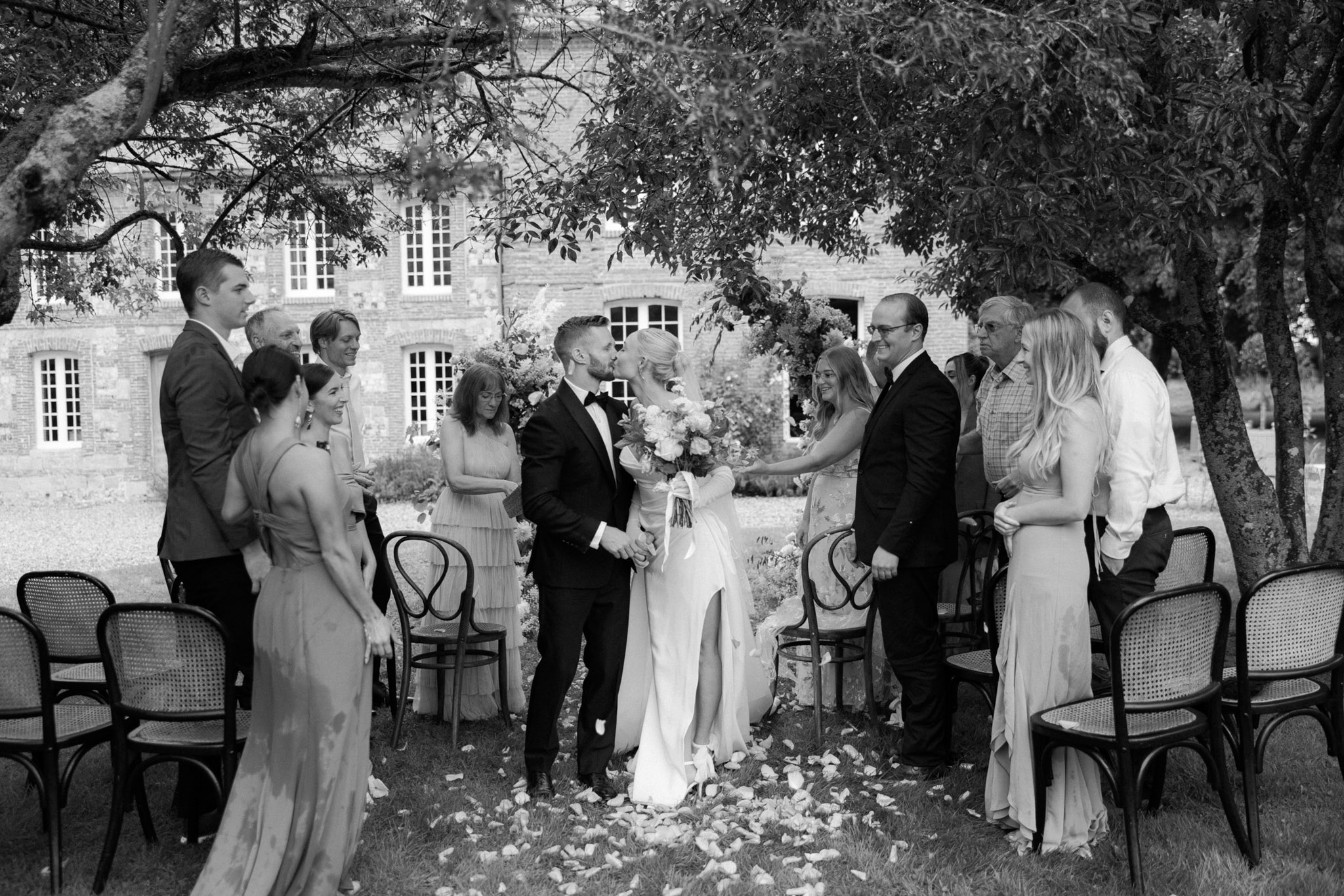 This black-and-white image captures the recessional moment of an outdoor wedding ceremony, with the bride and groom sharing a kiss at the end of the aisle while approximately ten guests standing on either side applaud and reach out to congratulate them. The ceremony takes place in the grounds of a large brick manor or château building visible in the background, with guests seated on classic bentwood cane chairs arranged in rows. The bride wears a long-sleeved fitted gown with a leg slit and holds a full bouquet with ribbon streamers, while the groom is dressed in a dark tuxedo with a bow tie. Flower petals are scattered across the grass aisle, and a floral arch is partially visible behind the couple. The image is a wide, medium-distance shot with soft contrast and fine detail throughout.