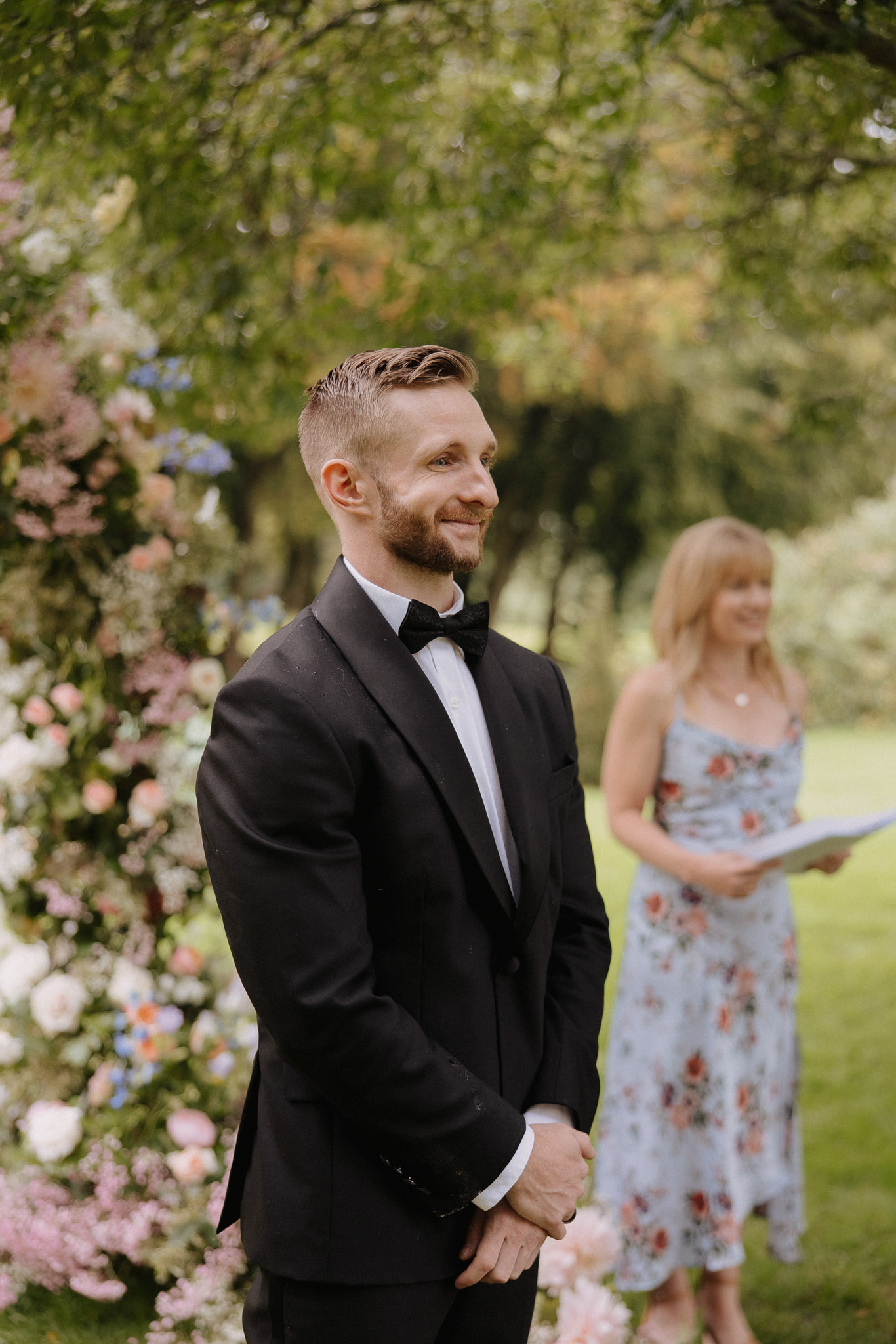 An outdoor wedding ceremony captured in a portrait shot, showing the groom standing at the altar watching the bride's approach. The groom wears a black tuxedo with a white dress shirt and black bow tie, hands clasped in front of him, smiling. To his left is a large floral arch decorated with blush pink astilbe, white garden roses, peach ranunculus, blue hydrangeas, and soft pink gypsophila against lush greenery. In the background, slightly out of focus, stands the officiant — a woman in a light blue floral-print midi dress holding a ceremony booklet. The setting is a garden lawn with mature trees providing a green backdrop, suggesting a classic outdoor garden ceremony aesthetic.